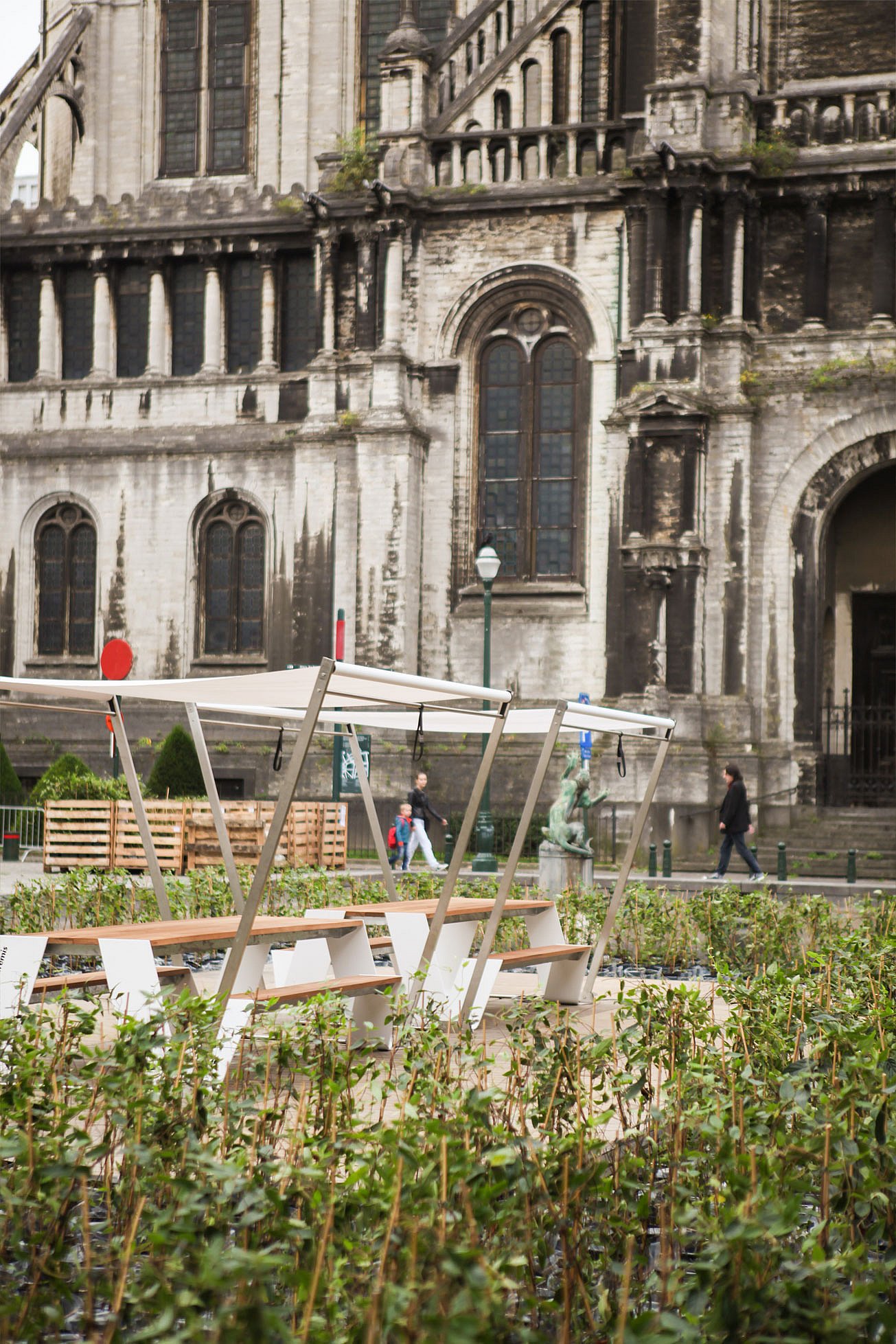Seitenansicht von zwei Hopper-Picknicktischen mit weißen Farbtönen, die sich über dichte Bepflanzung erheben, mit der verwitterten Kirchenfassade und Fußgängern dahinter.