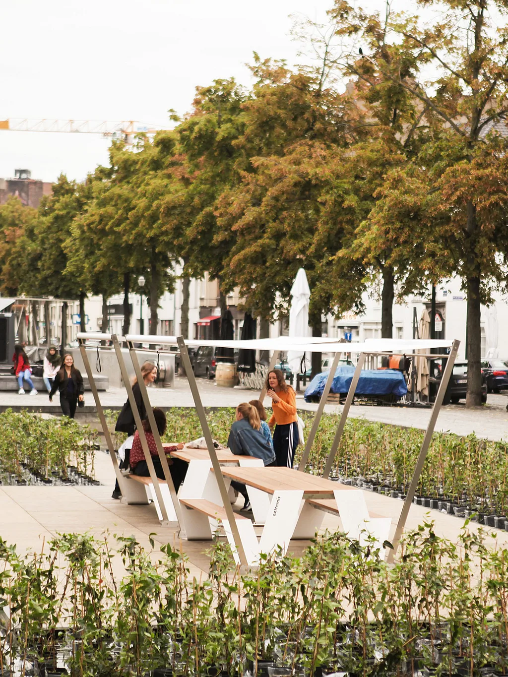 Hopper picnic table with white shade canopy on a wooden platform surrounded by rows of young plants in Brussels, with several people seated and tree-lined city streets in the background.