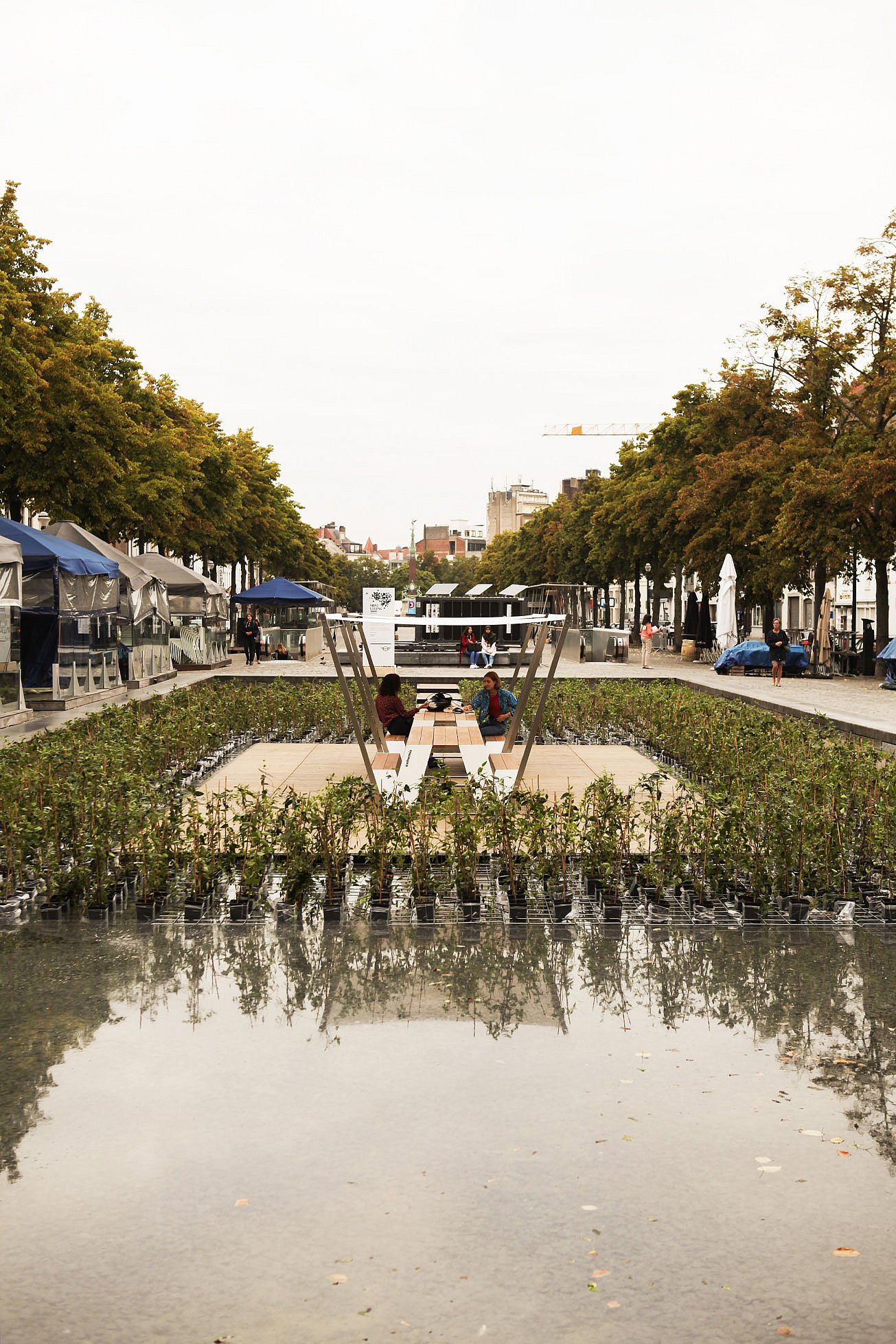 Weitblick über ein reflektierendes Wasserspiel auf einen zentralen Hopper-Picknicktisch unter weißen Farbtönen, umgeben von bepflanzten Beeten und einem von Bäumen gesäumten öffentlichen Platz.