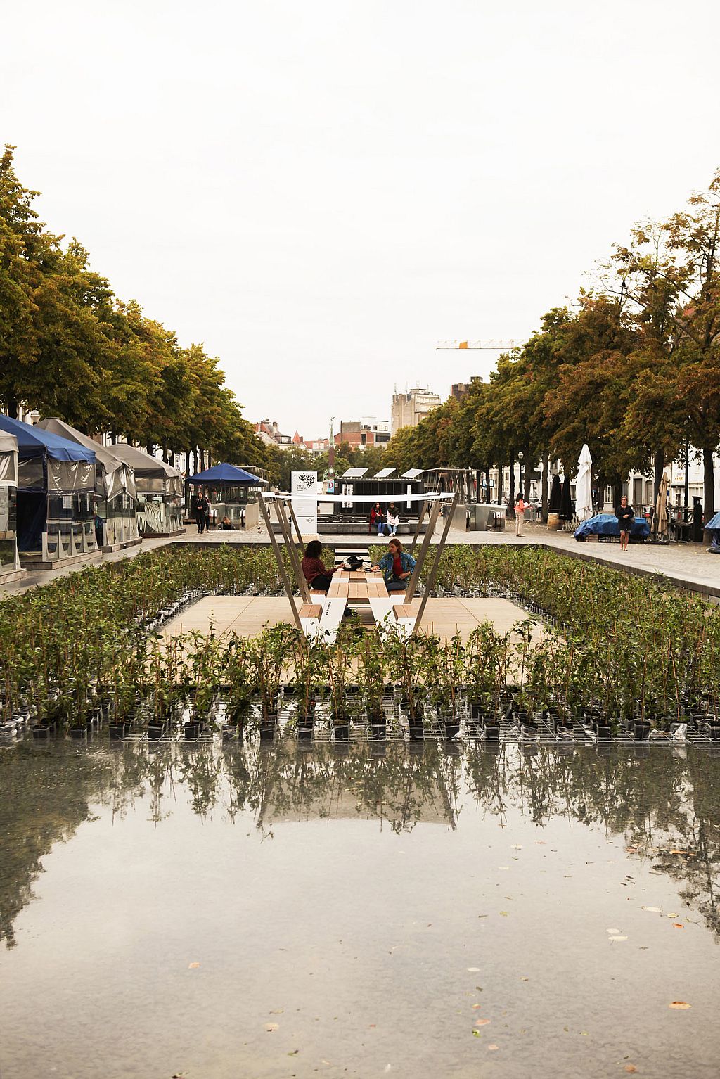 Weitblick über ein reflektierendes Wasserspiel auf einen zentralen Hopper-Picknicktisch unter weißen Farbtönen, umgeben von bepflanzten Beeten und einem von Bäumen gesäumten öffentlichen Platz.