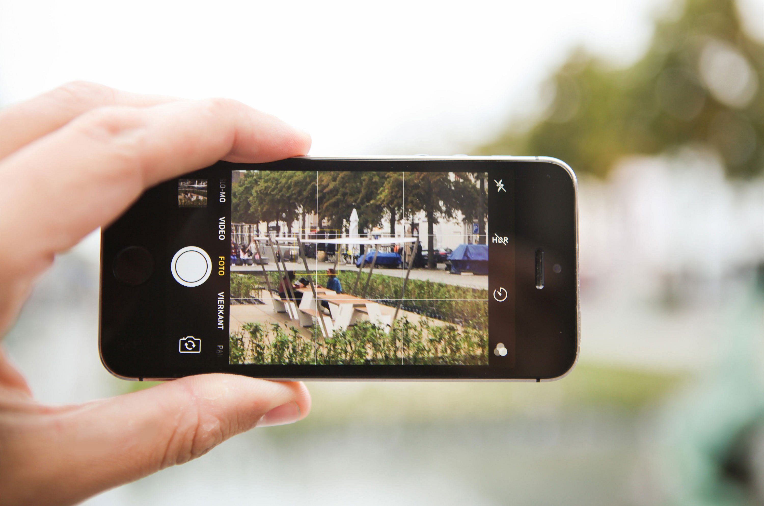 Hand hält ein Smartphone, während sie die schattige Hopper-Picknicktischinstallation fotografiert, wobei die Szene auf dem Telefonbildschirm eingerahmt und im Hintergrund unscharf ist.