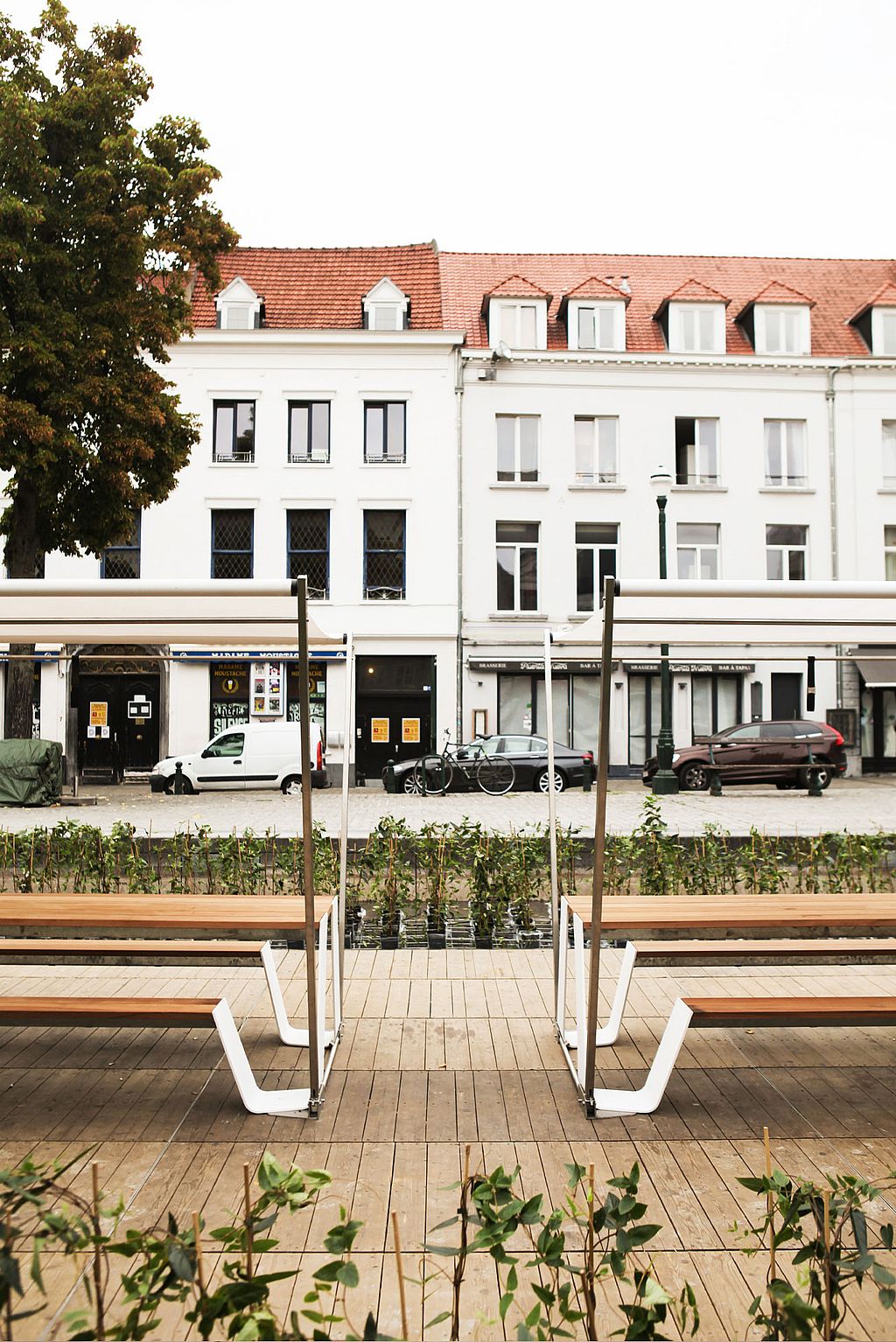 Rückansicht von zwei Hopper-Picknicktischen unter weißen Jalousien auf einer Holzterrasse, gegenüber einer Reihe weißer Stadthäuser auf der anderen Seite des Platzes.
