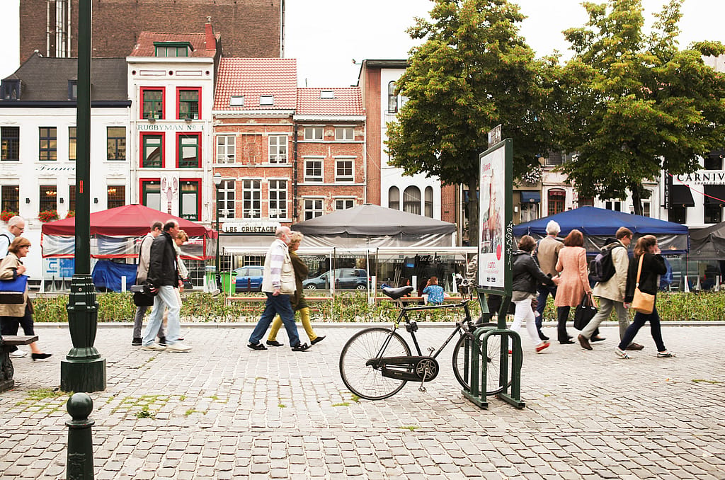 Fußgänger, die über einen Kopfsteinpflasterplatz an einem geparkten Fahrrad und einer Werbetafel vorbeigehen, hinter ihnen sind die bepflanzte MINI Living-Installation und schattige Sitzgelegenheiten sichtbar.