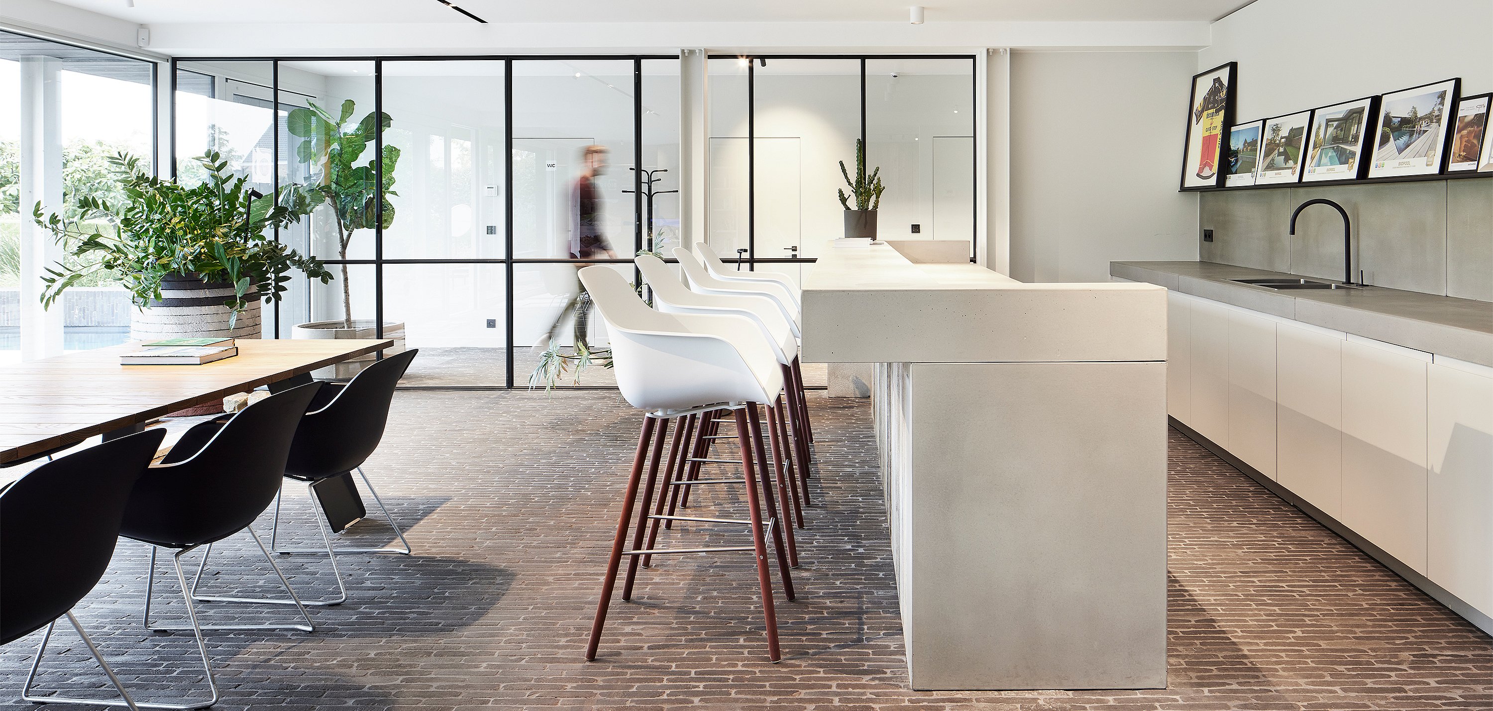 Helle Büroküche mit einer großen Betoninsel, weißen hohen Kapitänsstühlen mit Holzbeinen, schwarzen Esszimmerstühlen und Glastrennwänden mit schwarzem Rahmen.