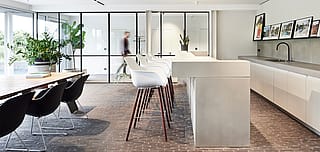 Helle Büroküche mit einer großen Betoninsel, weißen hohen Kapitänsstühlen mit Holzbeinen, schwarzen Esszimmerstühlen und Glastrennwänden mit schwarzem Rahmen.