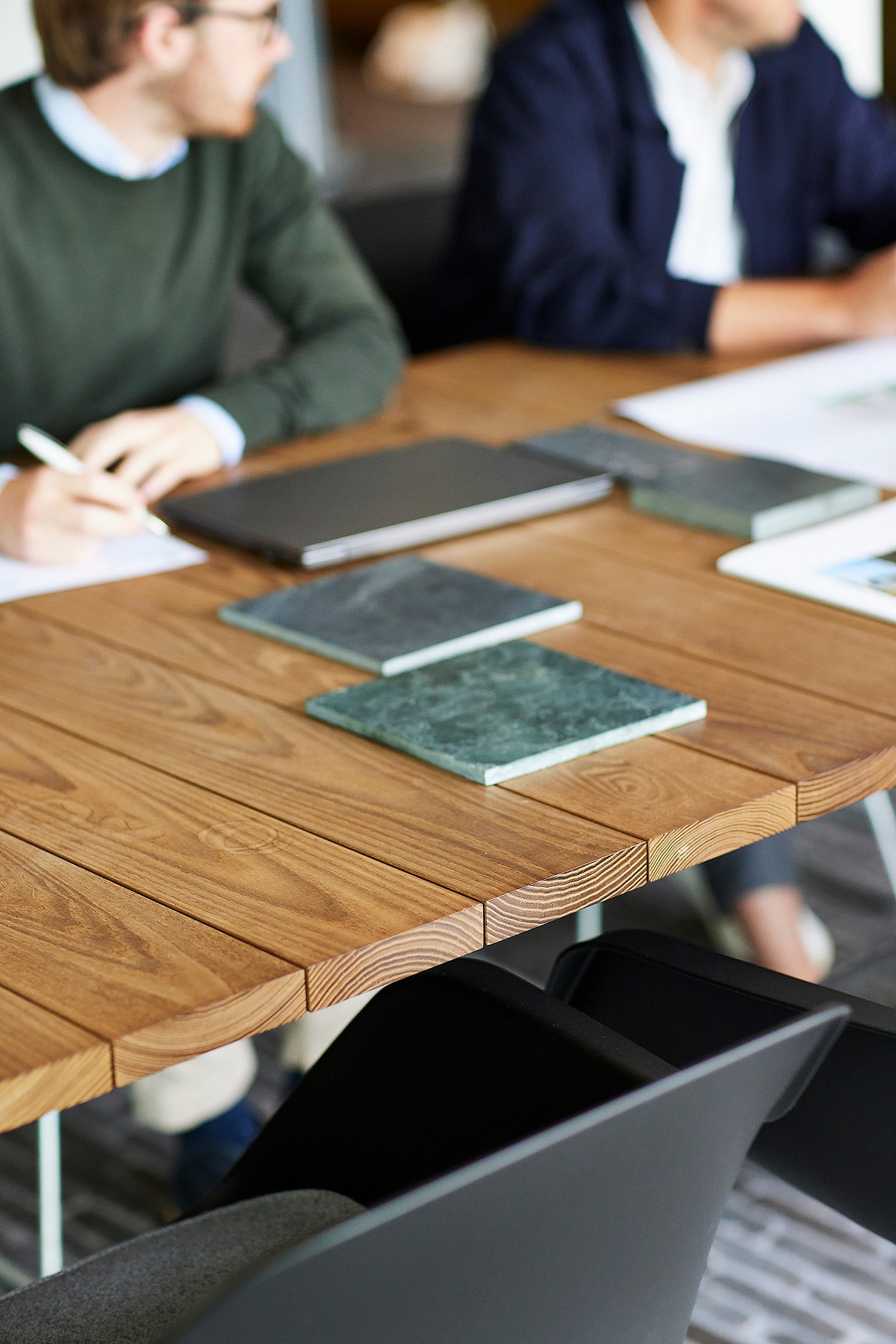 Besprechungstisch aus Holz mit ausgelegten Steinmustern, Notizbüchern und Dokumenten, während Kollegen im Hintergrund sitzen und reden.