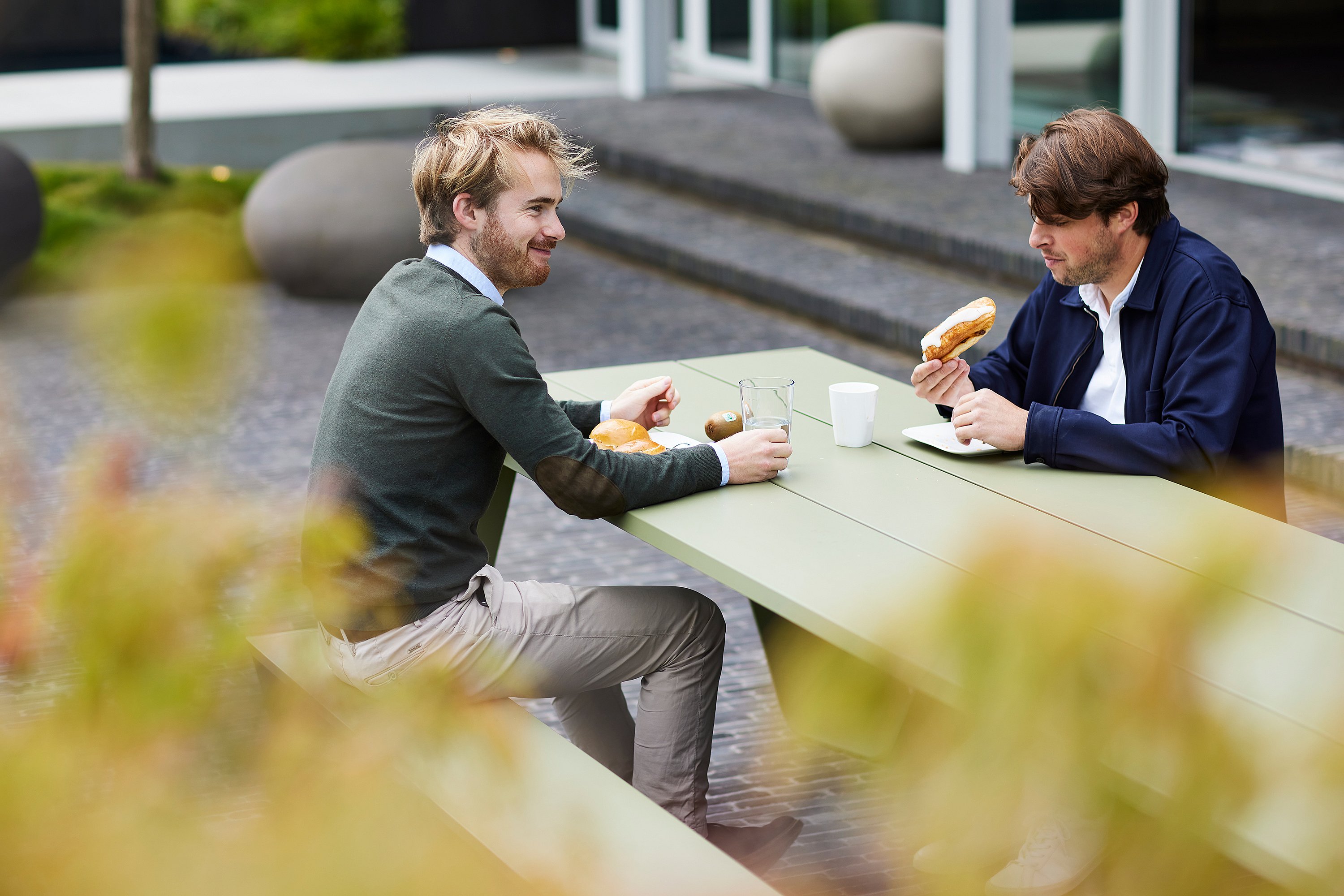 Zwei Kollegen essen an einem grünen Trichtertisch im Freien auf der Terrasse zu Mittag, um den herum die Bürotreppe und die Bepflanzung sanft verschwommen sind.