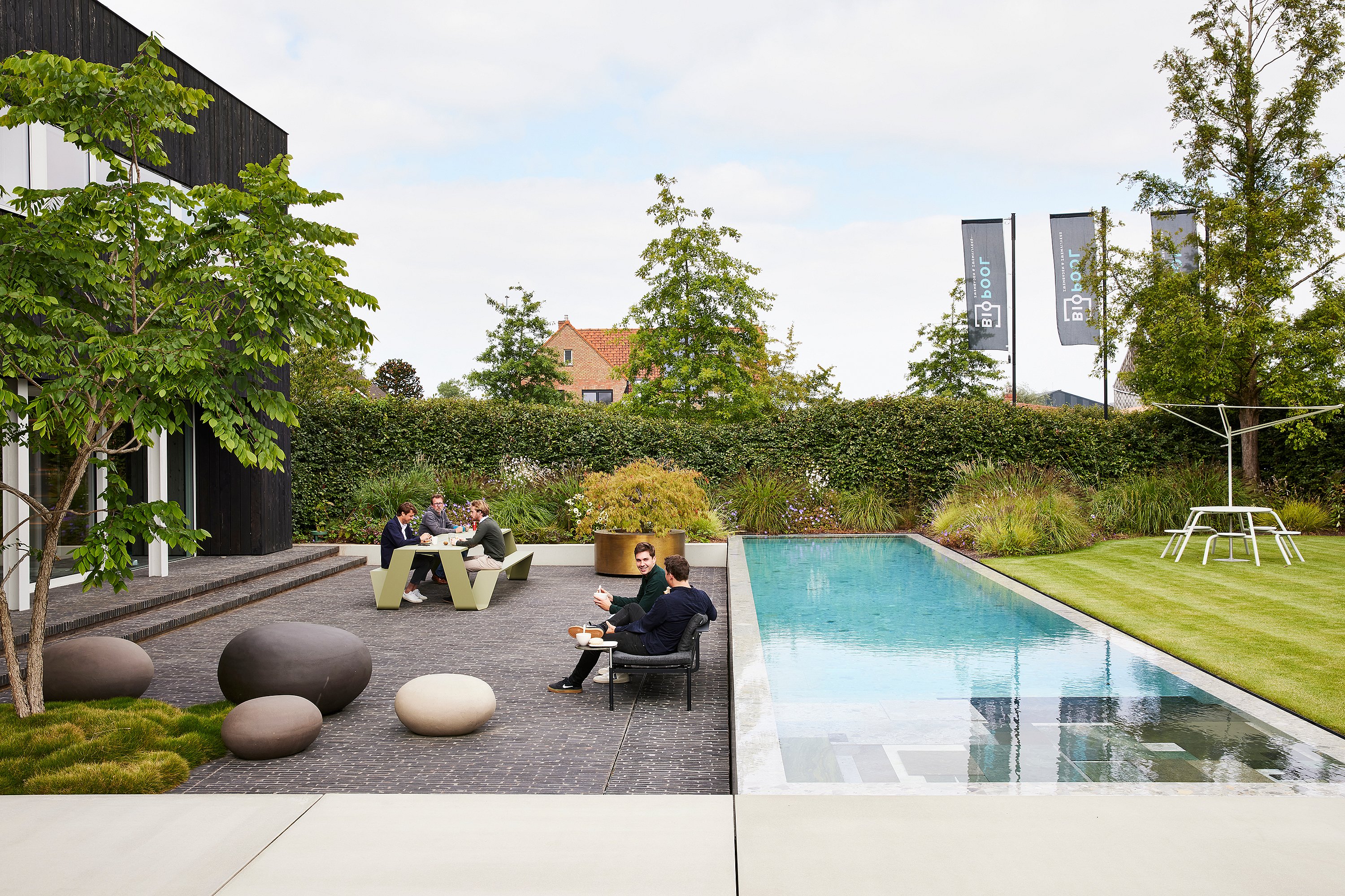Gartenterrasse mit langem Swimmingpool, wo Kollegen auf einem schwarzen Outdoor-Sofa und an einem grünen Trichtertisch sitzen, mit einem Virus-Picknicktisch und Akazienschatten auf dem Rasen sichtbar.