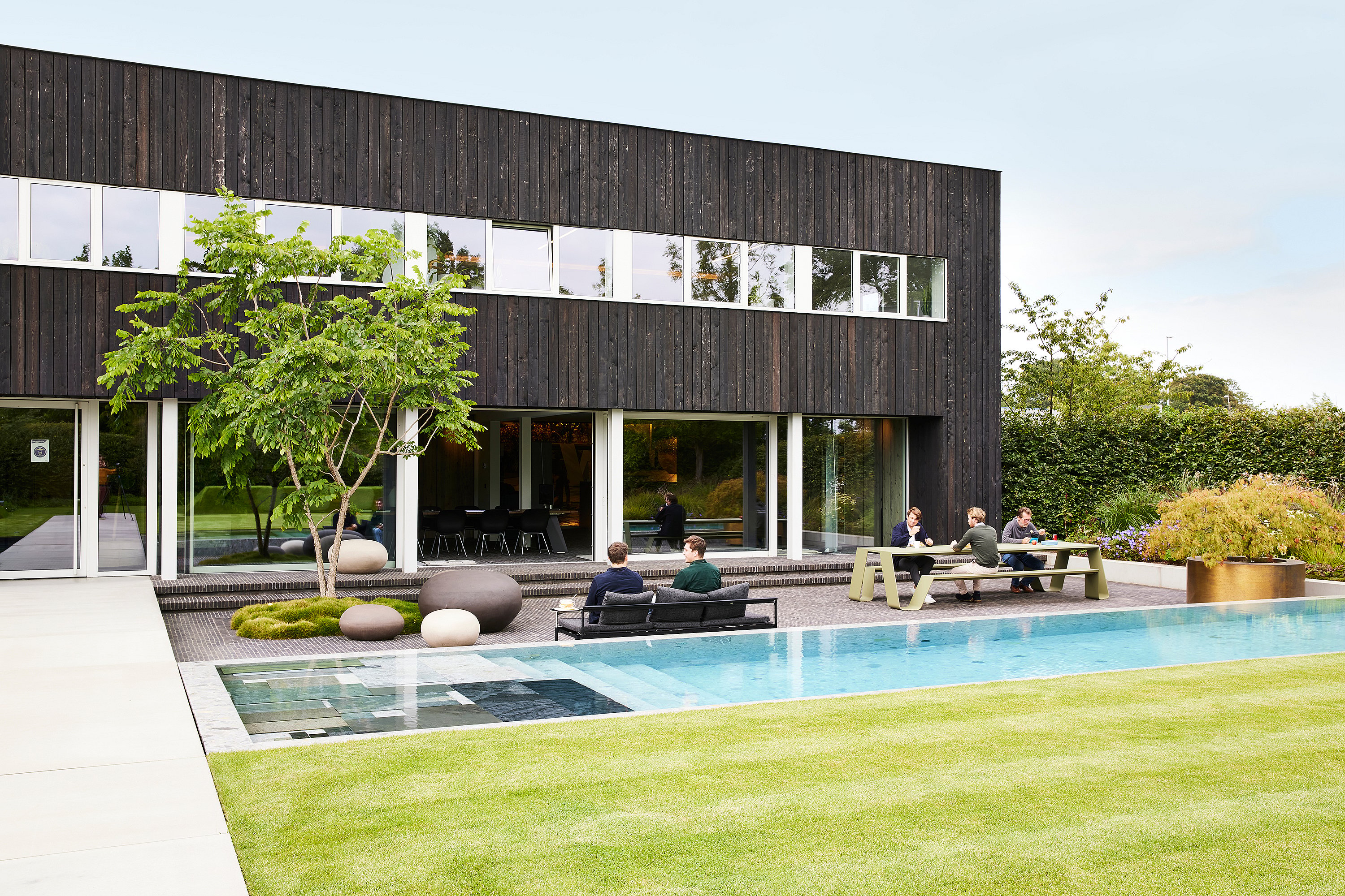 Immeuble de bureaux moderne en bois noir avec une longue piscine, où les collègues s’assoient sur un sol noir + canapé luna et à une table en entonnoir vert sur la terrasse à côté d’une pelouse soigneusement coupée.