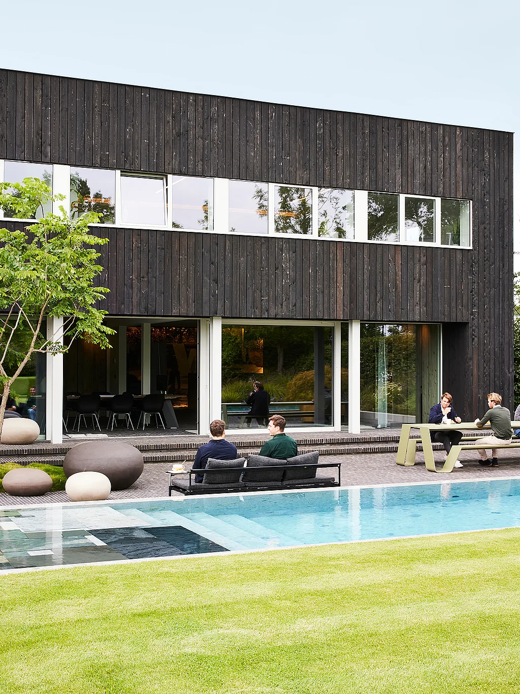 Modern zwart houten kantoorgebouw met een lang zwembad, waar collega's op een zwarte sol + luna bank zitten en aan een groene trechter tafel op het terras naast een netjes bijgesneden gazon.