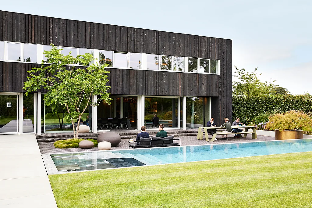 Immeuble de bureaux moderne en bois noir avec une longue piscine, où les collègues s’assoient sur un sol noir + canapé luna et à une table en entonnoir vert sur la terrasse à côté d’une pelouse soigneusement coupée.