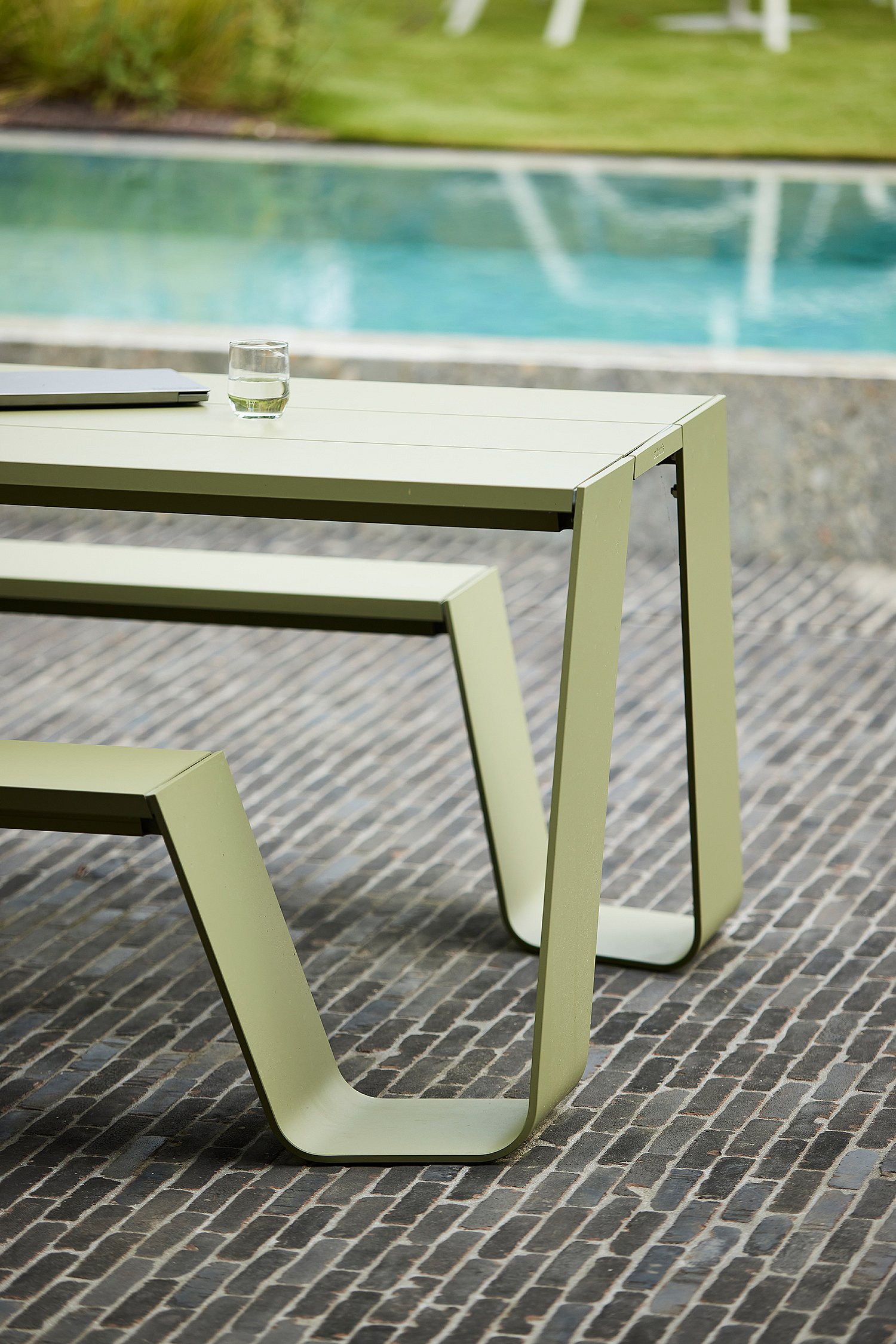 Nahaufnahme eines grünen Trichtertisches und einer grünen Bank im Freien neben dem Pool, mit einem Glas Wasser und einem geschlossenen Laptop auf der Tischplatte.