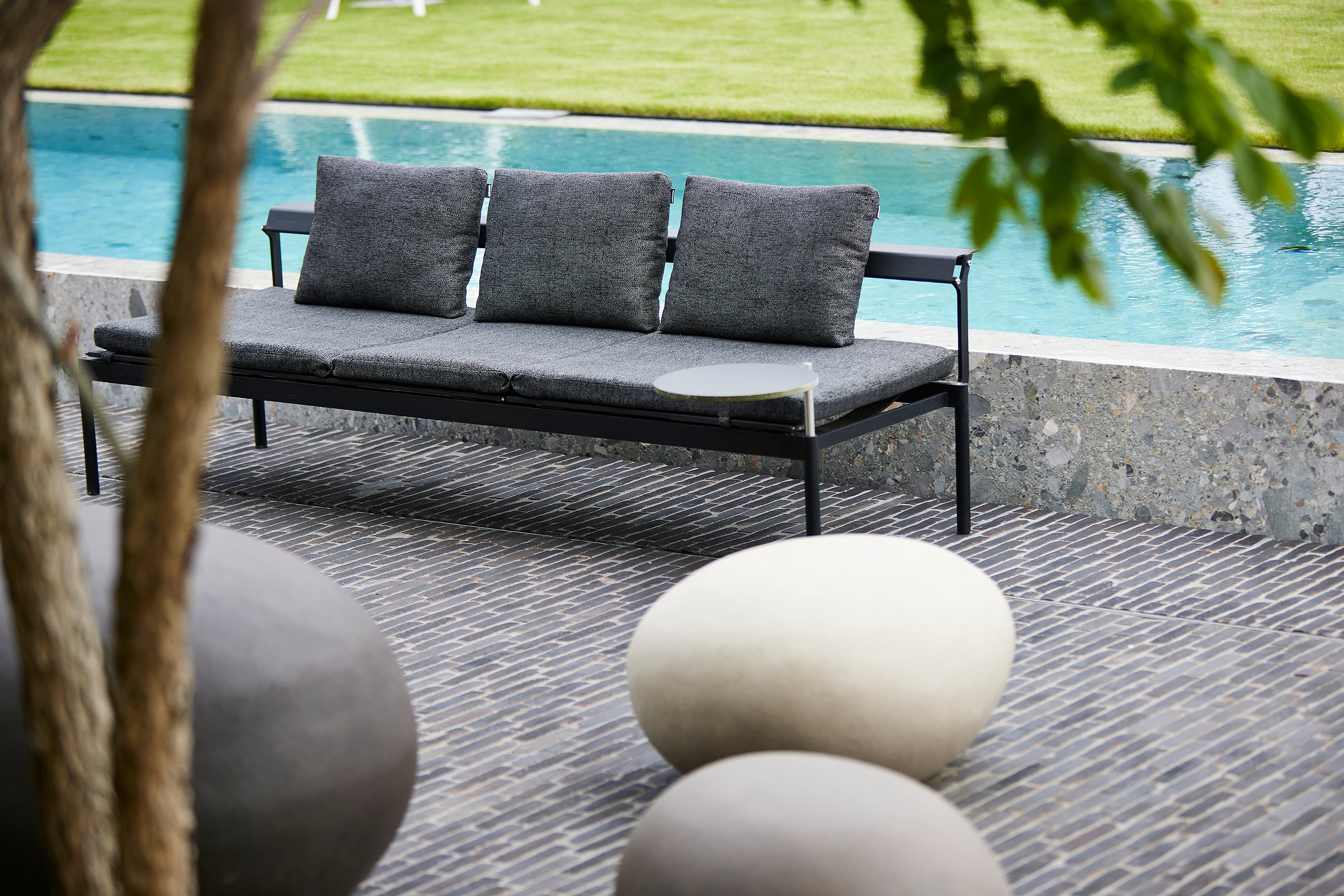Leeres schwarzes Sol & Luna-Sofa für den Außenbereich mit drei grauen Extremkissen auf der Rückseite neben dem Pool, eingerahmt von glatten, runden Gartensteinen im Vordergrund.