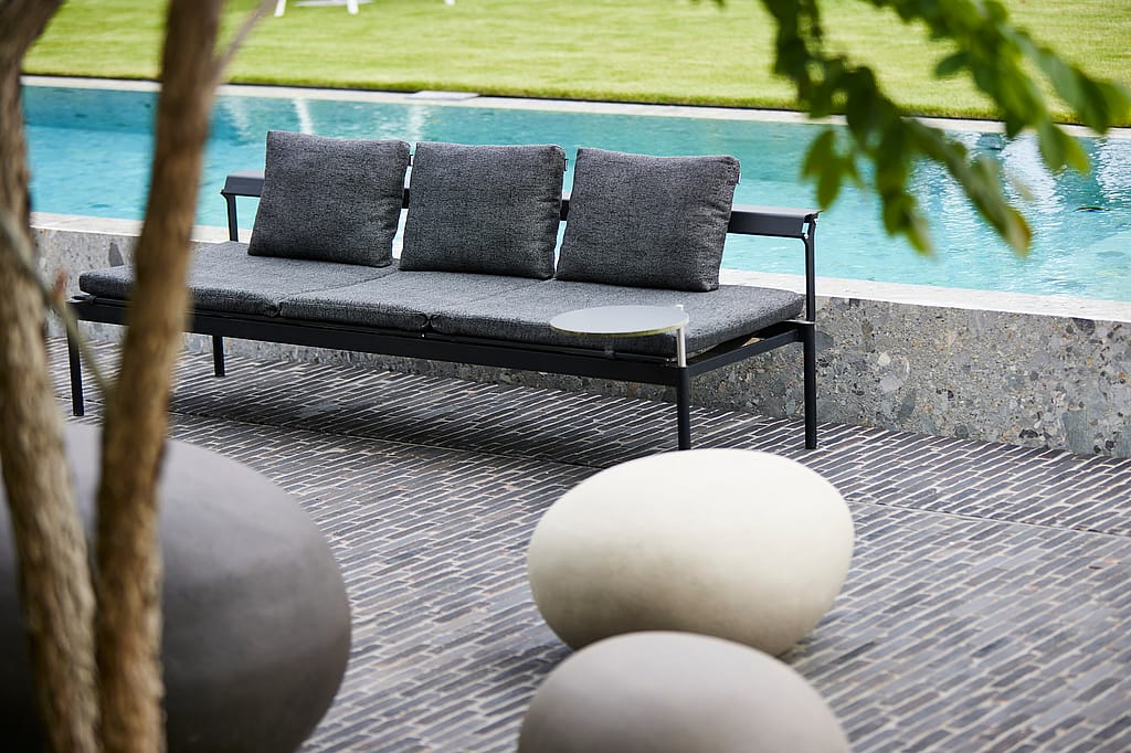 Leeres schwarzes Sol & Luna-Sofa für den Außenbereich mit drei grauen Extremkissen auf der Rückseite neben dem Pool, eingerahmt von glatten, runden Gartensteinen im Vordergrund.