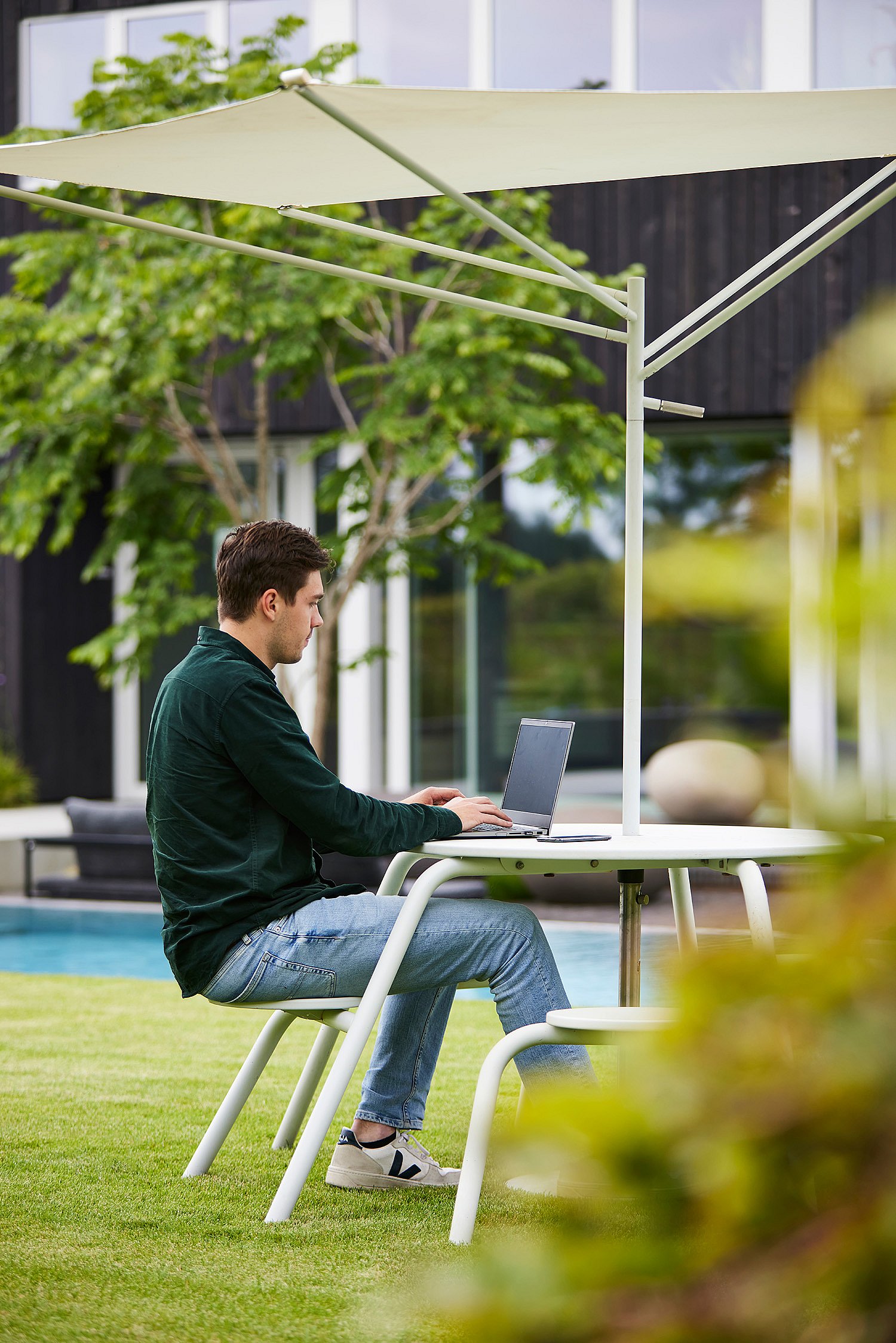 Man working on a laptop at a white outdoor table with integrated shade on a lawn beside a modern house.