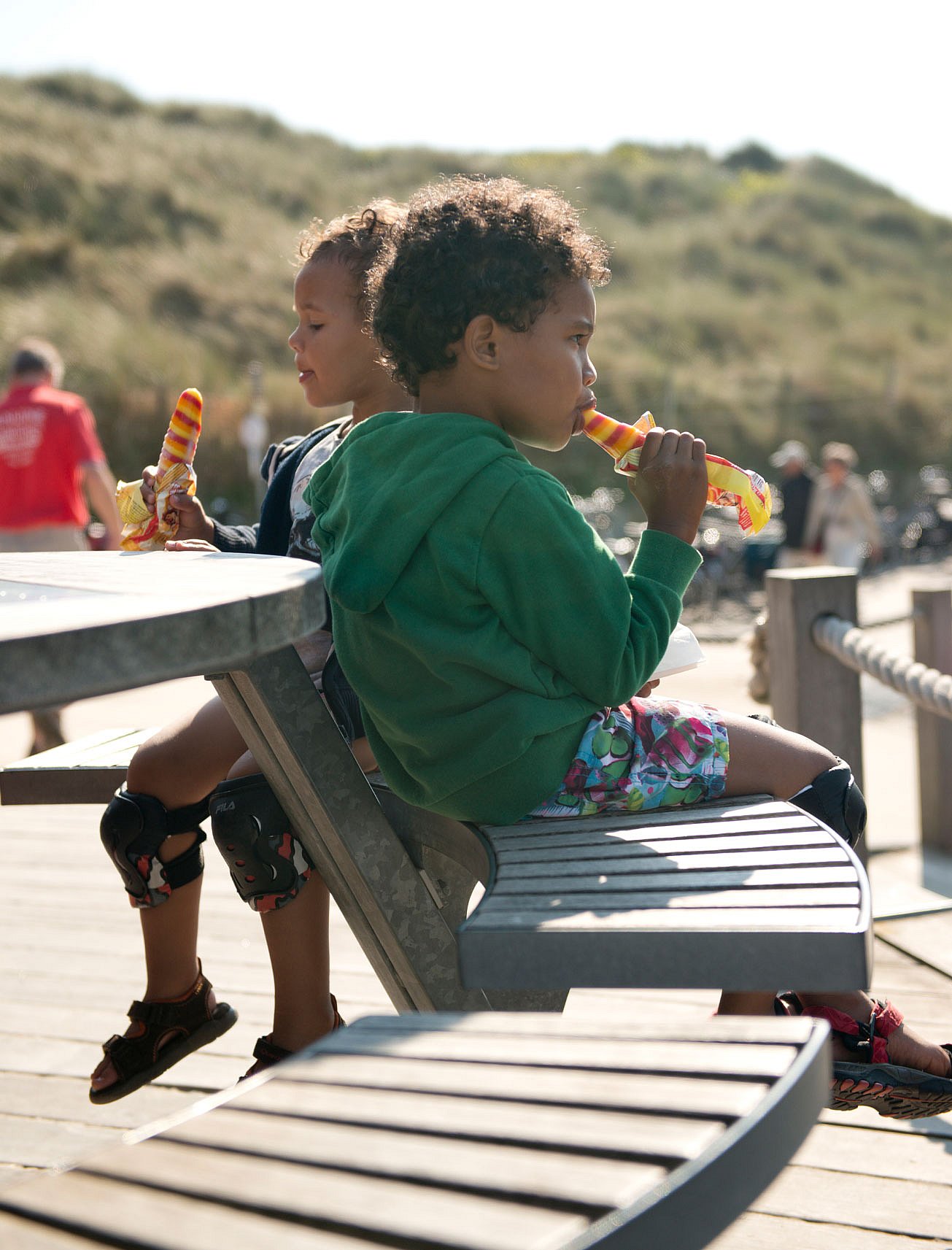  Deux enfants assis dos à dos sur le banc incurvé d'une table ronde extérieure, dégustant des glaces à l'eau rayées sur la promenade du bord de mer.  Vue panoramique du bar de plage Twins en bois avec ses terrasses étagées, ses tables de pique-nique blanches, ses herbes des dunes et la plage qui s'étend à côté.