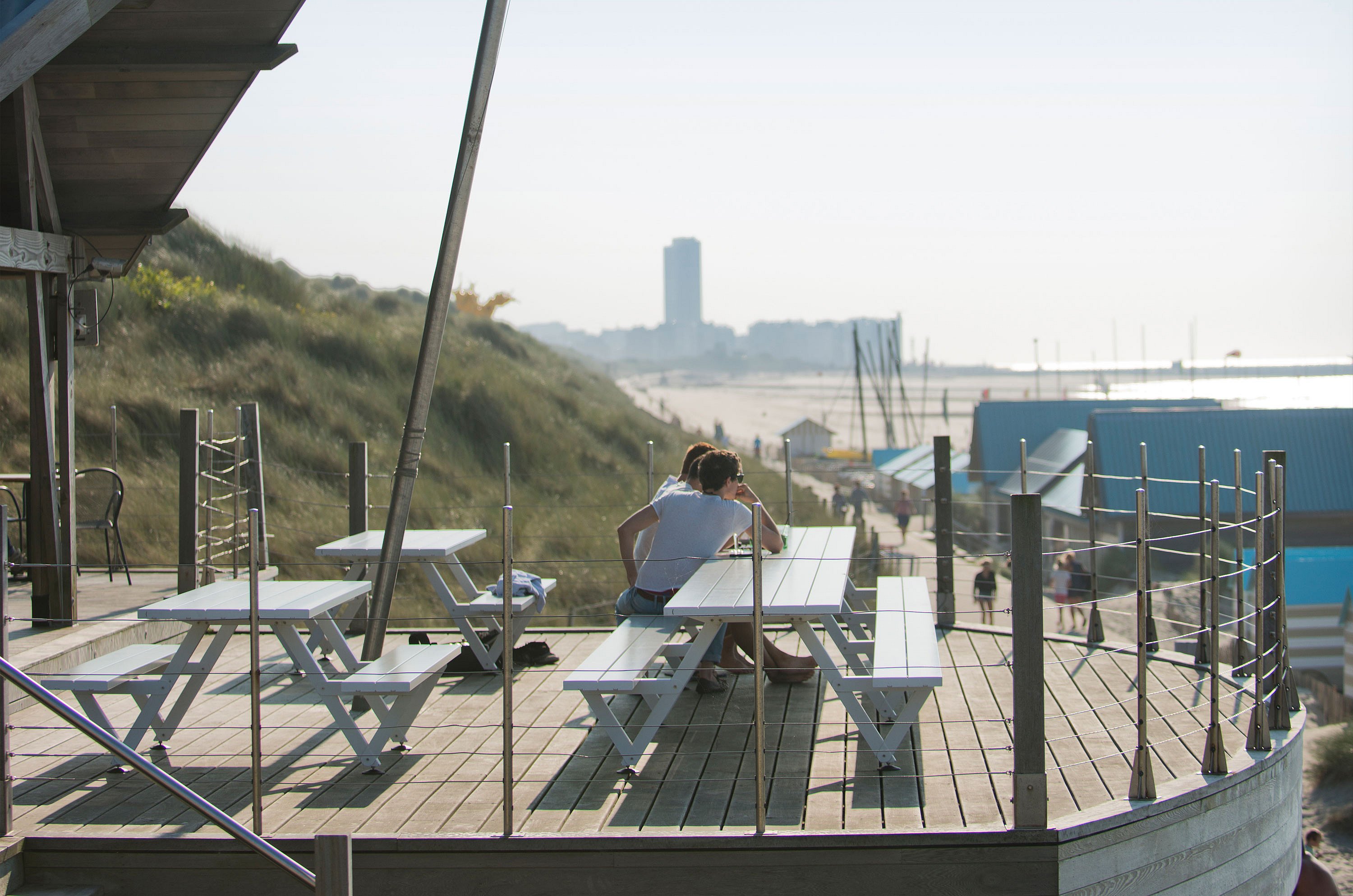 Witte Marina picknicktafels op een gebogen houten strandterras, met twee mensen naast elkaar en duinen, strandhuisjes en de zee in de verte.
