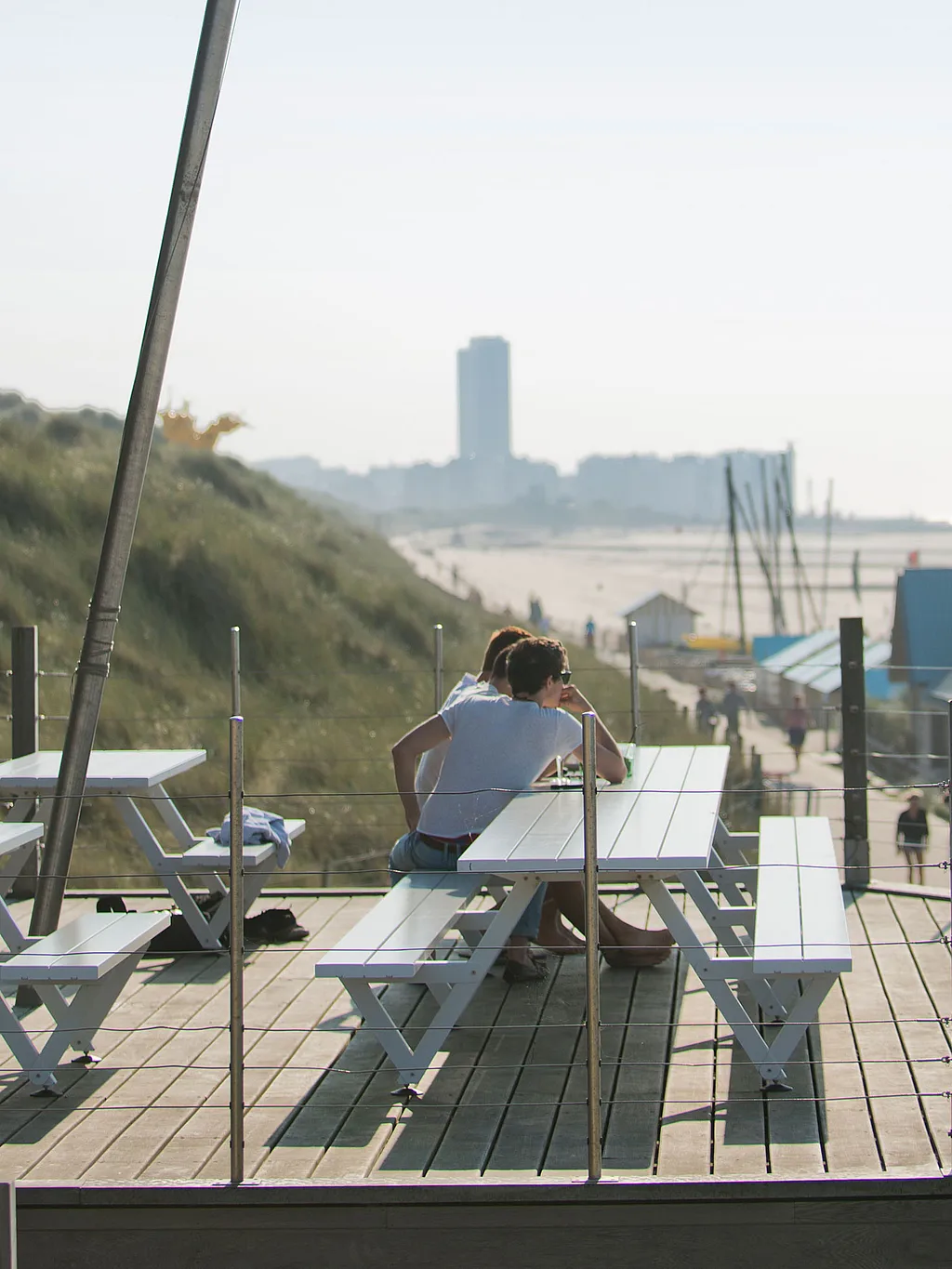 Witte Marina picknicktafels op een gebogen houten strandterras, met twee mensen naast elkaar en duinen, strandhuisjes en de zee in de verte.