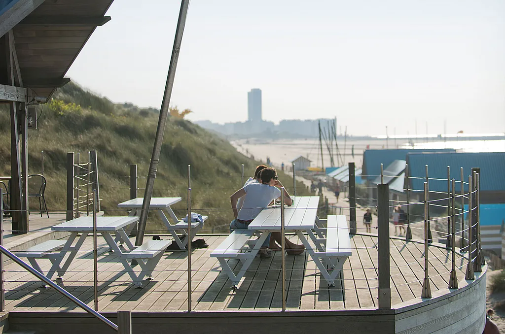 White Marina picnic tables on a curved wooden beach terrace, with two people seated together and dunes, beach huts, and the sea in the distance.