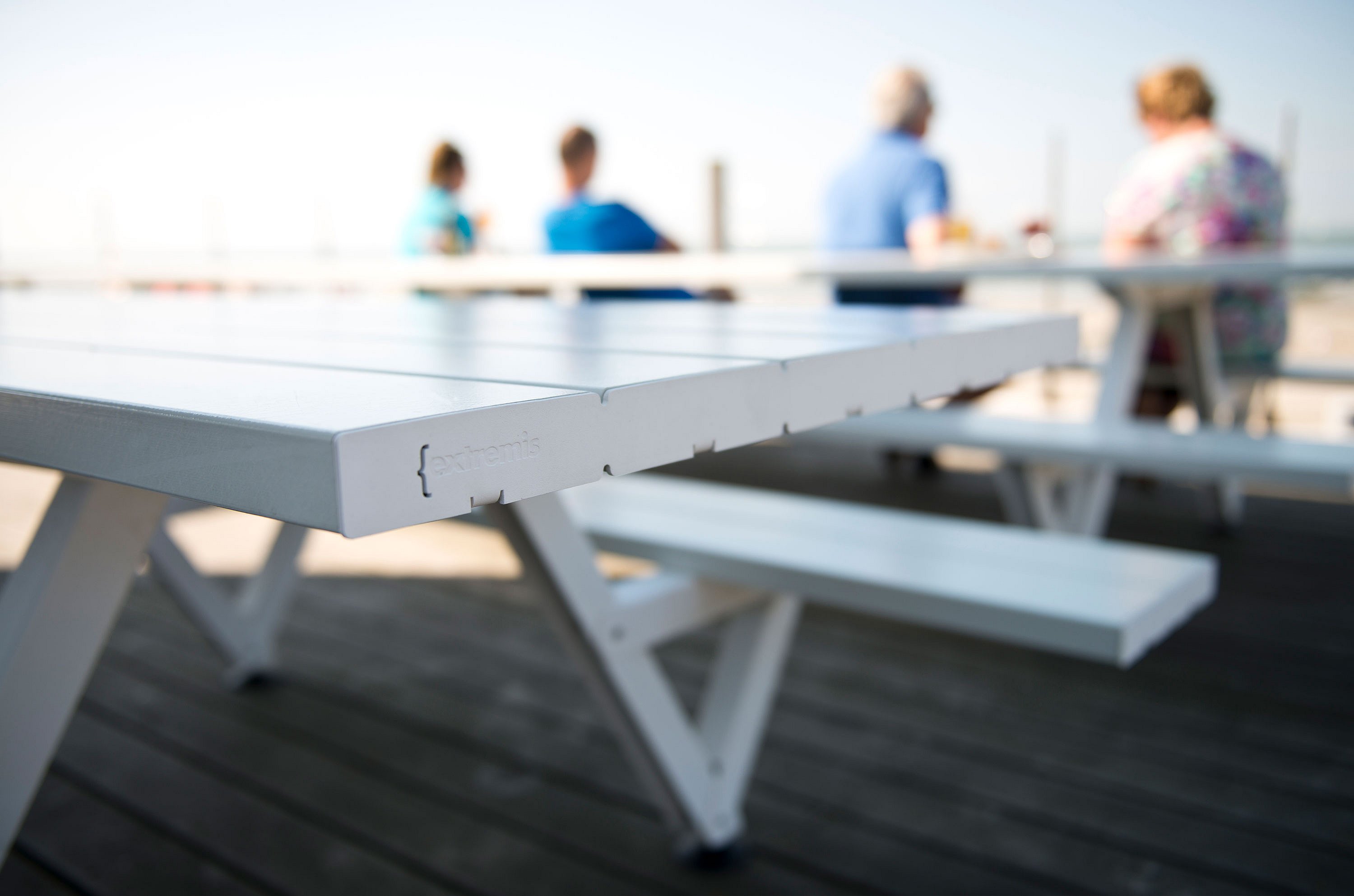 Gros plan sur le coin d'une table de pique-nique blanche avec le logo Extremis, avec des baigneurs assis à une autre table, flous en arrière-plan.