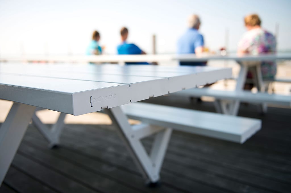 Gros plan sur le coin d'une table de pique-nique blanche avec le logo Extremis, avec des baigneurs assis à une autre table, flous en arrière-plan.