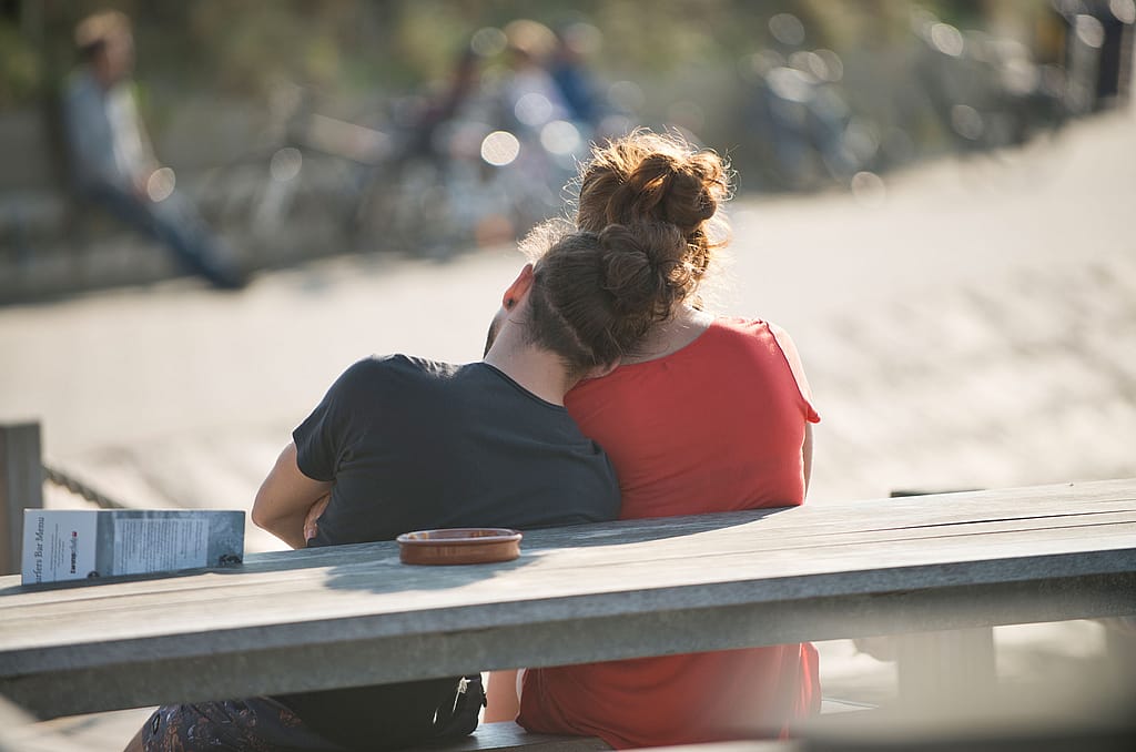 Deux personnes assises côte à côte sur un banc de plage en bois, vues de dos sur un rivage de sable légèrement flou.