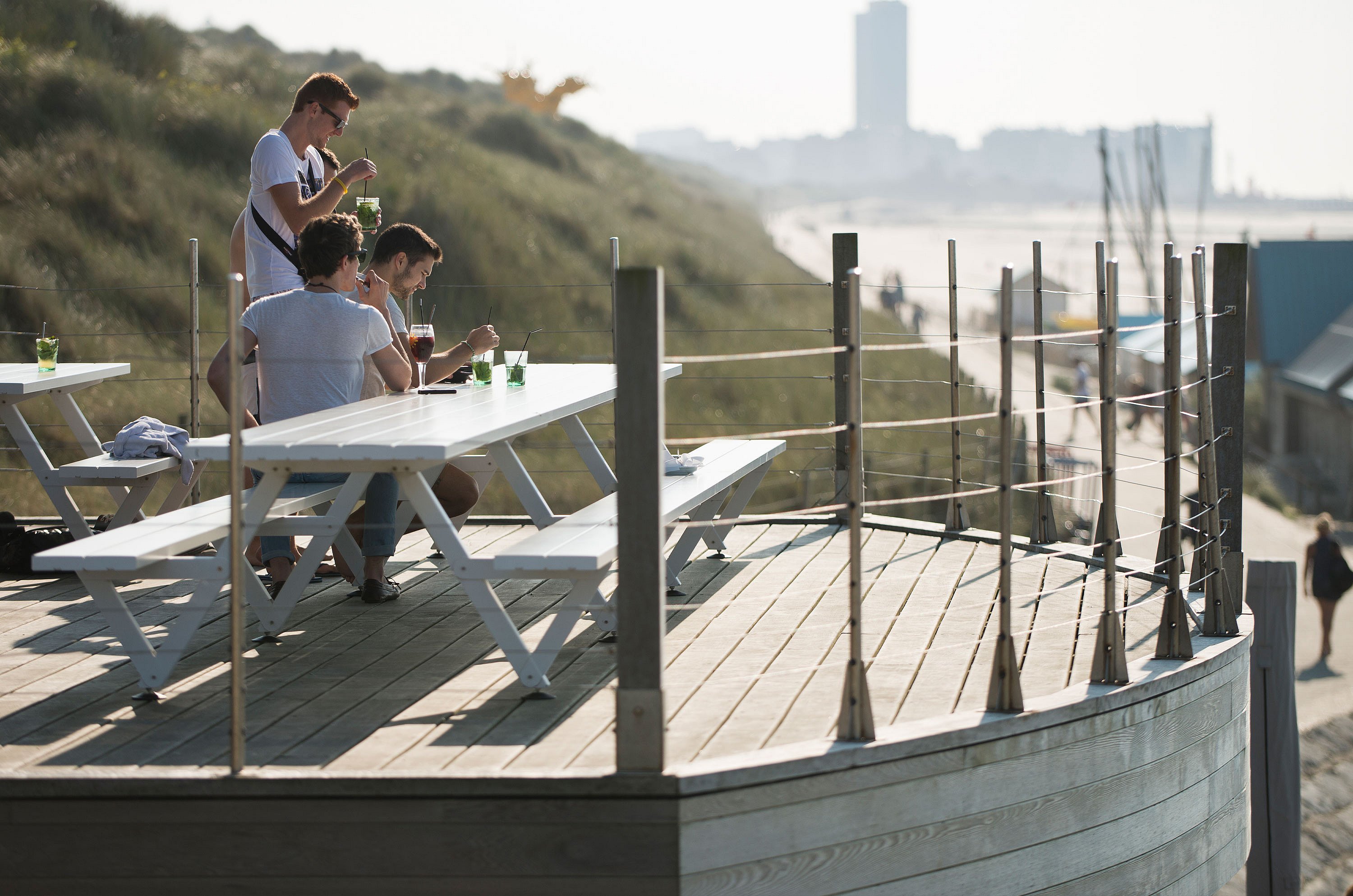 Trois amis prenant un verre à une table de pique-nique blanche Hopper sur une terrasse surélevée au-dessus de la plage, avec les dunes et le rivage en arrière-plan.