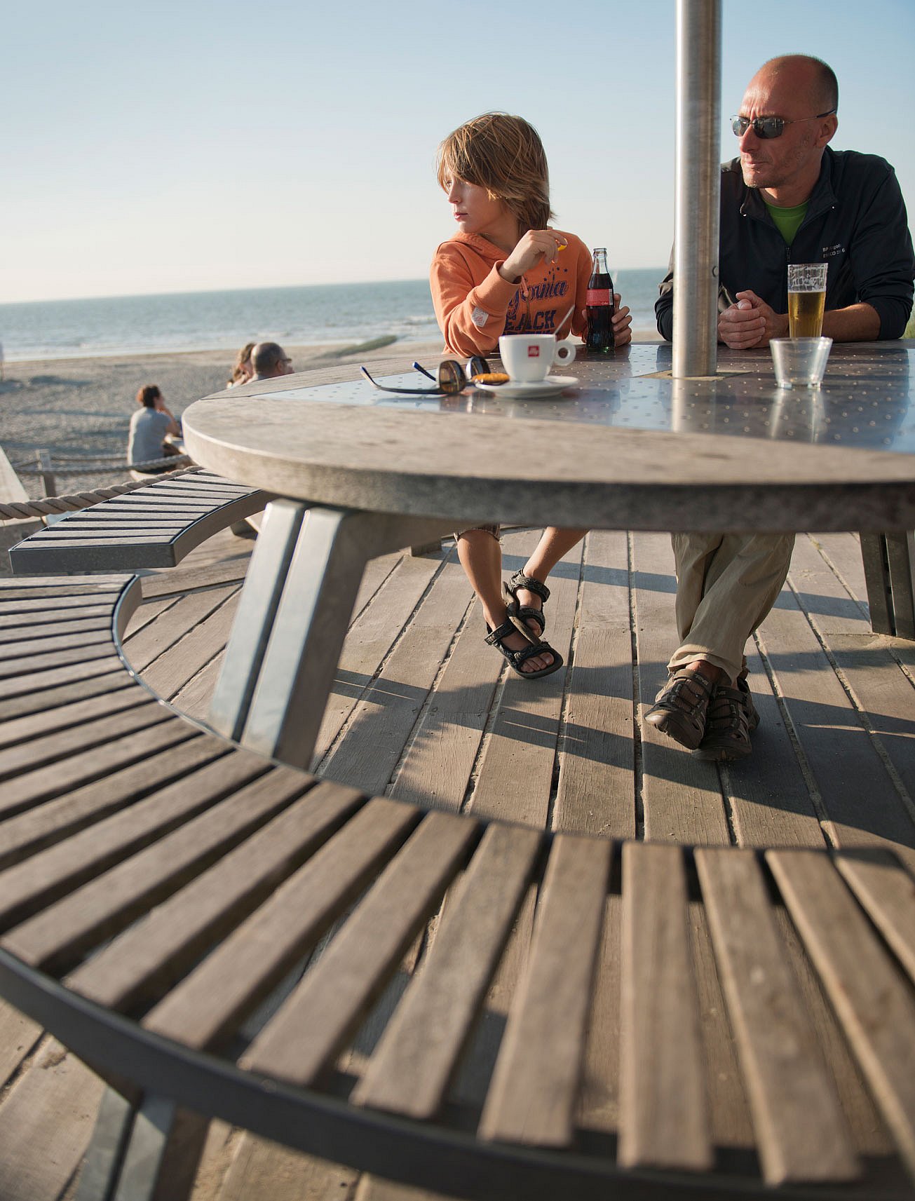   Un enfant et un homme assis à une table ronde extérieure avec des boissons, contemplant la plage et la mer sous le soleil de fin d'après-midi.