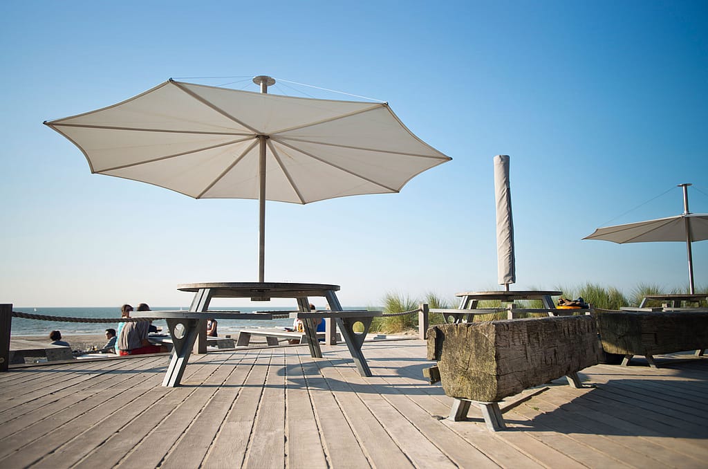 Table ronde d'extérieur sous un grand parasol blanc sur une terrasse en bois, avec des herbes des dunes, des bancs et la plage au loin.