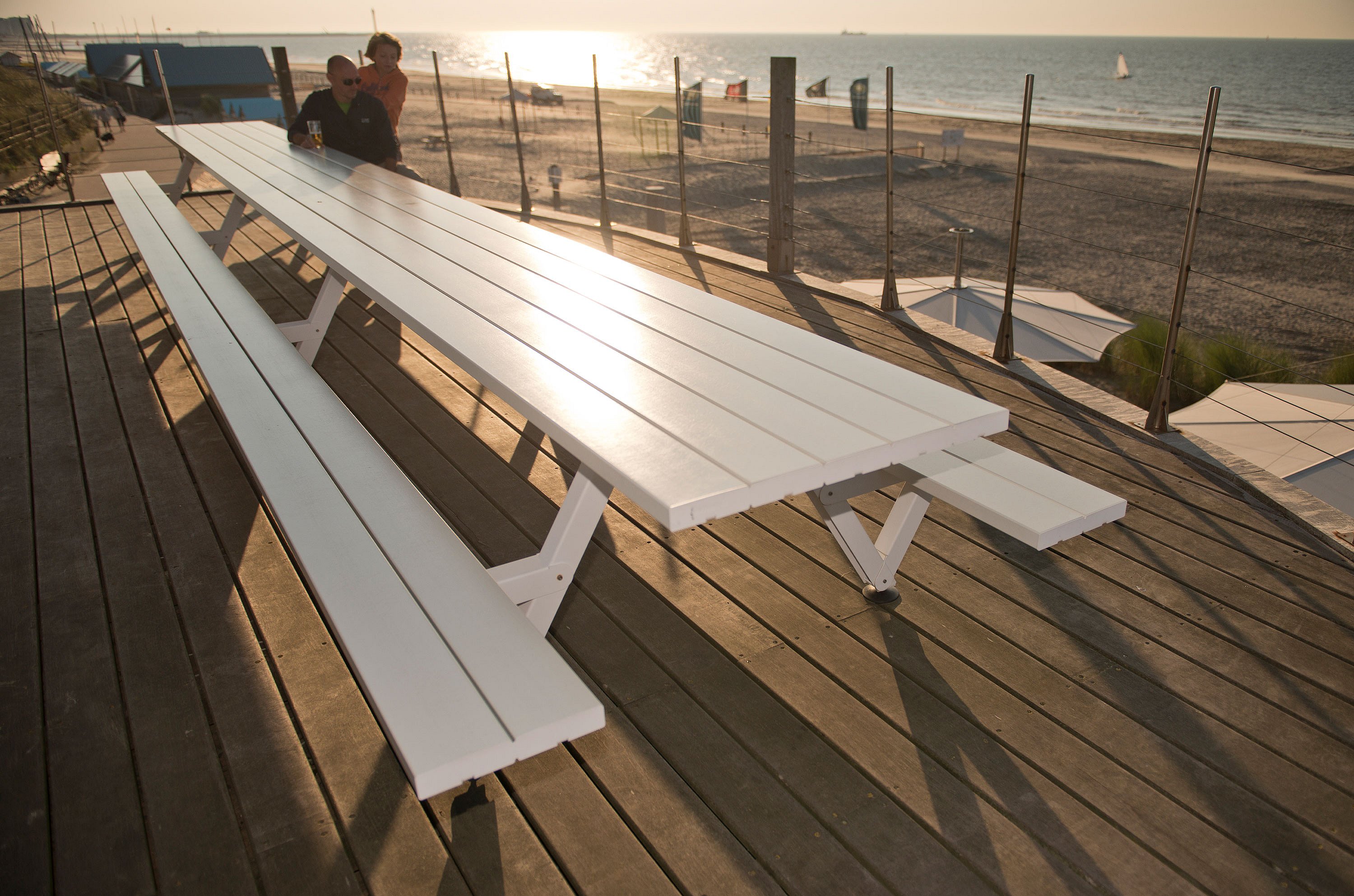   Longue table de pique-nique blanche Marina sur une terrasse en bois surplombant la plage, avec deux personnes à l'extrémité et la mer scintillante en arrière-plan.