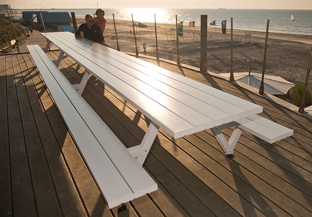 Longue table de pique-nique blanche Marina sur une terrasse en bois surplombant la plage, avec deux personnes à l'extrémité et la mer scintillante en arrière-plan.