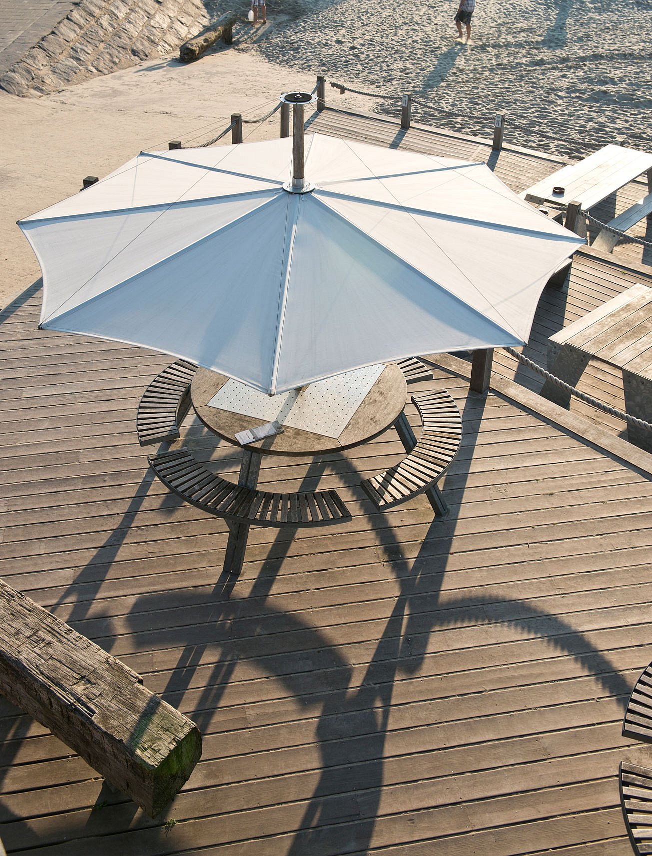   Vue aérienne d'une table ronde Gargantua avec des bancs en bois incurvés sous un grand parasol blanc sur une terrasse ensoleillée en bord de plage.