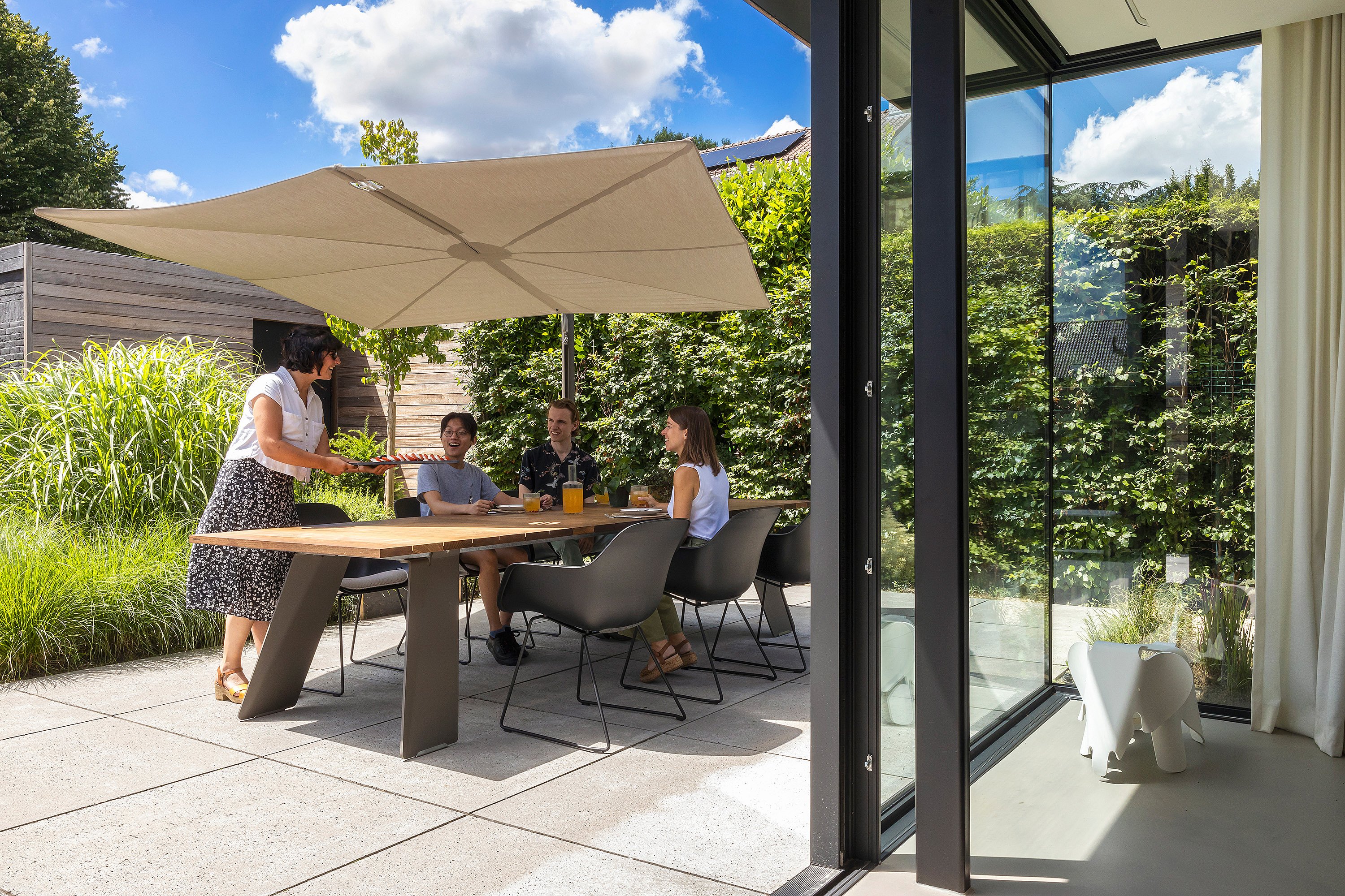Four people dining at a wooden outdoor table under a large beige Sensu parasol beside a modern glass-walled house.