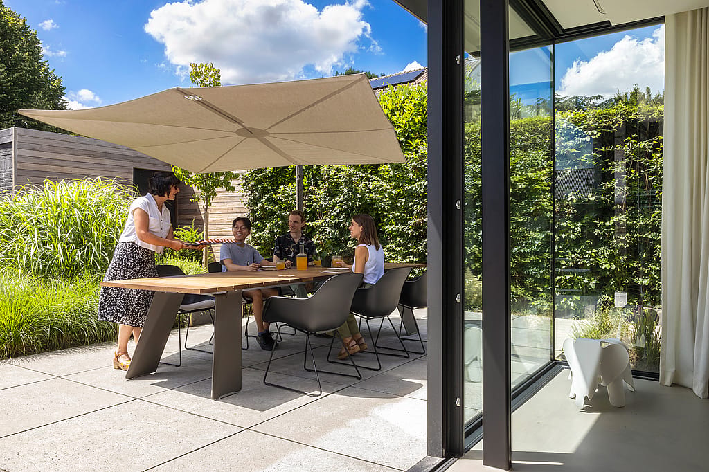 Four people dining at a wooden outdoor table under a large beige Sensu parasol beside a modern glass-walled house.