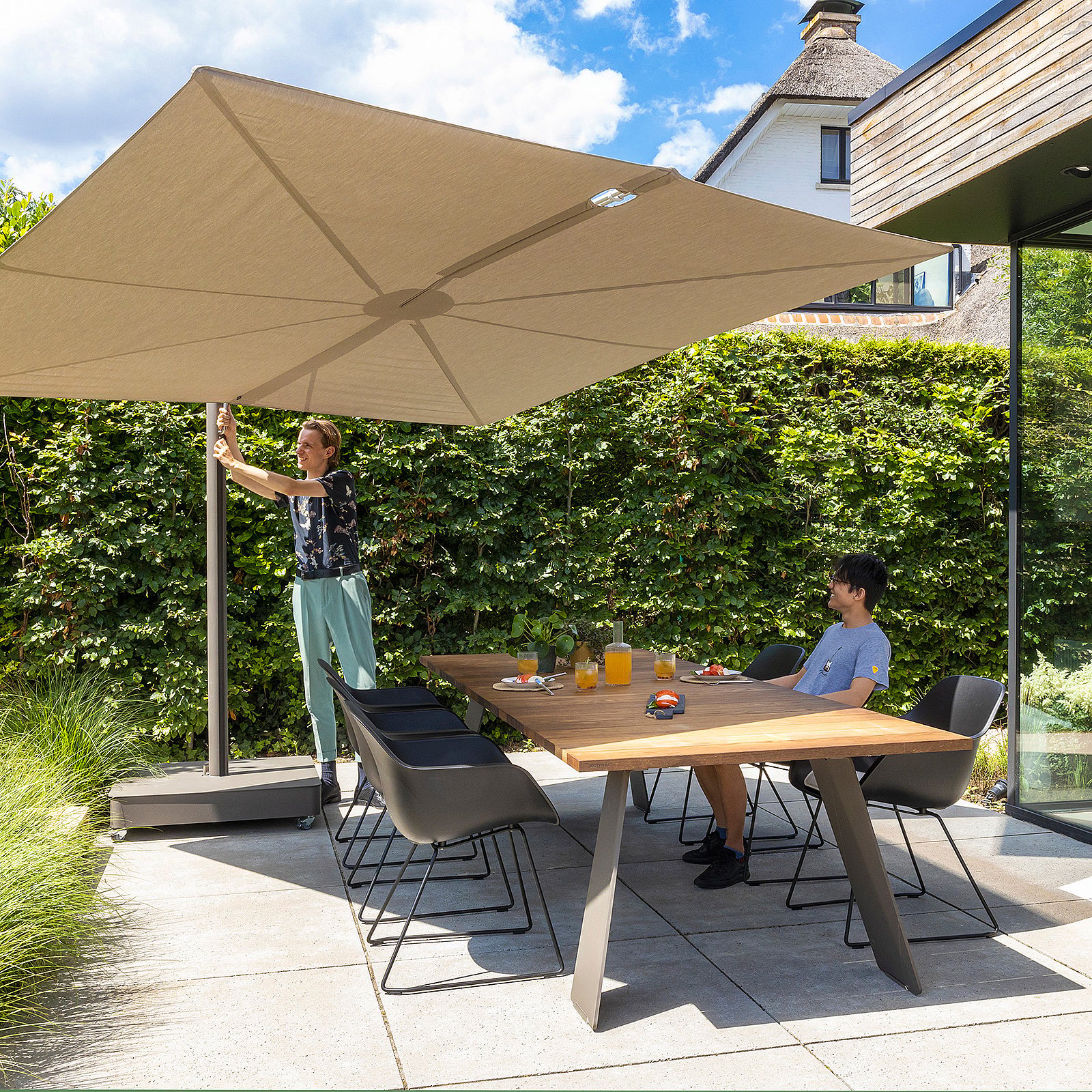 Person adjusting a large beige Sensu parasol beside a wooden outdoor dining table with black chairs and drinks in the sun.