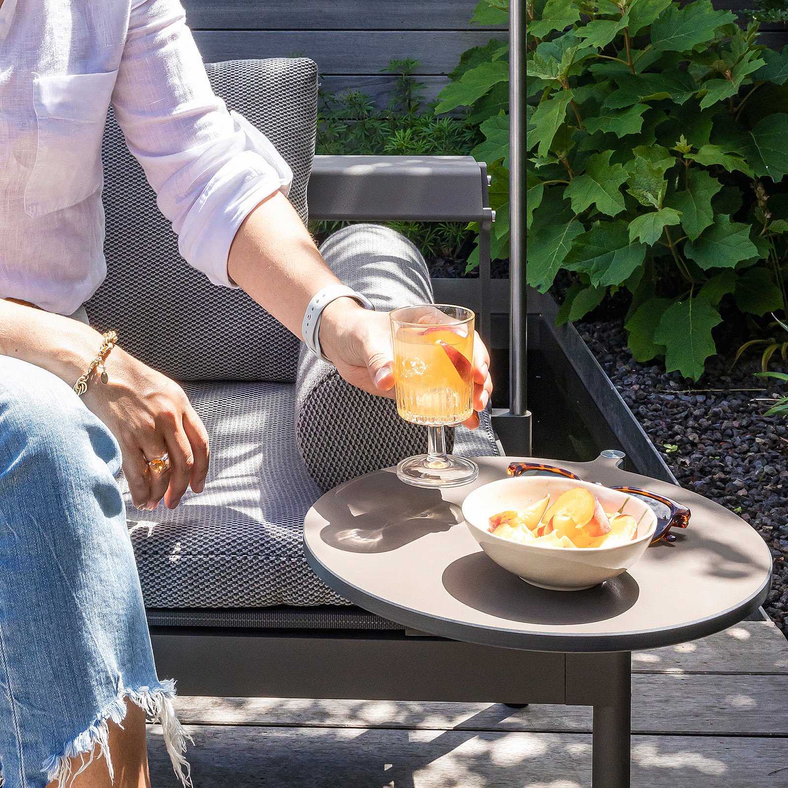 Close-up of a hand holding an orange drink above a round side table with a bowl of fruit beside a gray outdoor lounger.