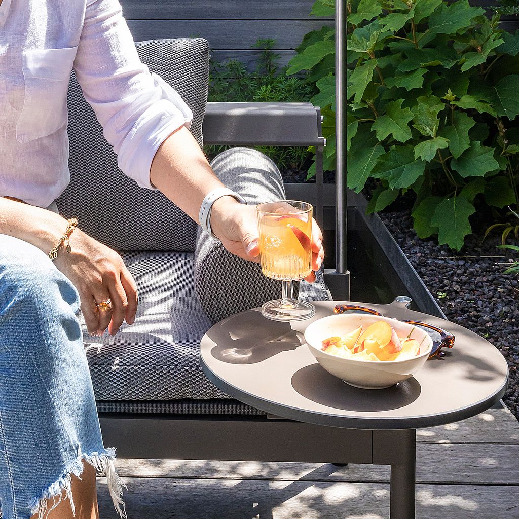 Close-up of a hand holding an orange drink above a round side table with a bowl of fruit beside a gray outdoor lounger.