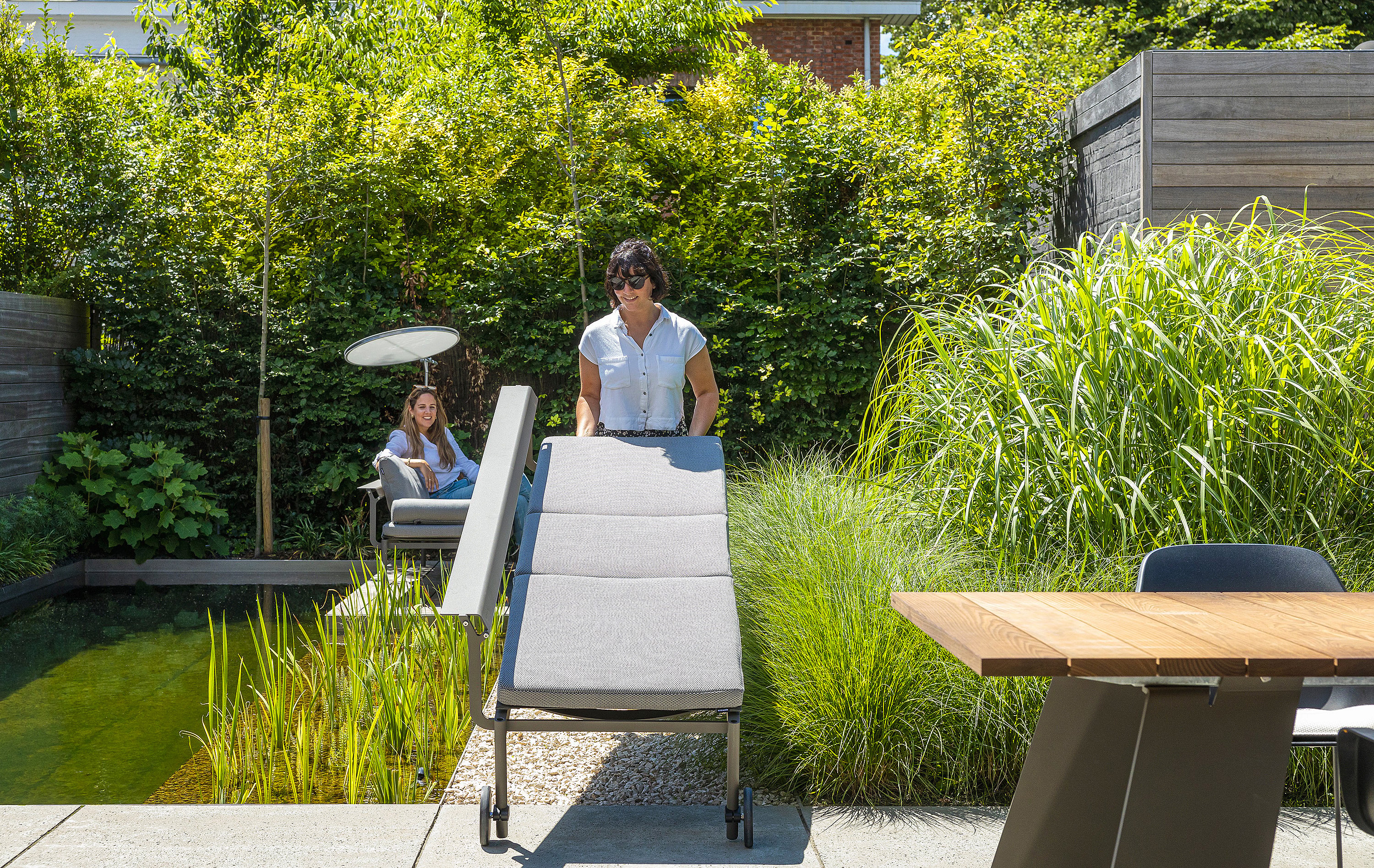 Woman rolling a gray Sol+Luna lounger with wheels across the terrace beside the natural pool, with another person seated in the background.