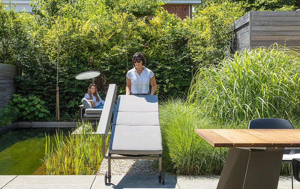 Woman rolling a gray Sol+Luna lounger with wheels across the terrace beside the natural pool, with another person seated in the background.