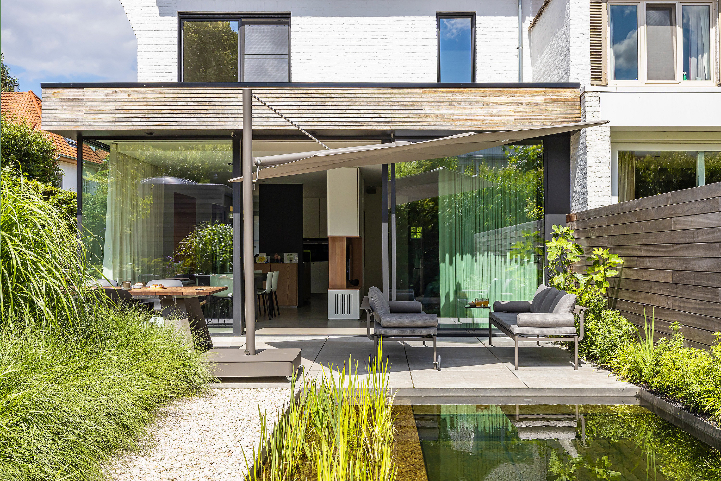 Modern rear terrace with gray Sol+Luna seating beside a natural pool, viewed from the garden toward a white house with large sliding glass doors and a shaded dining area.