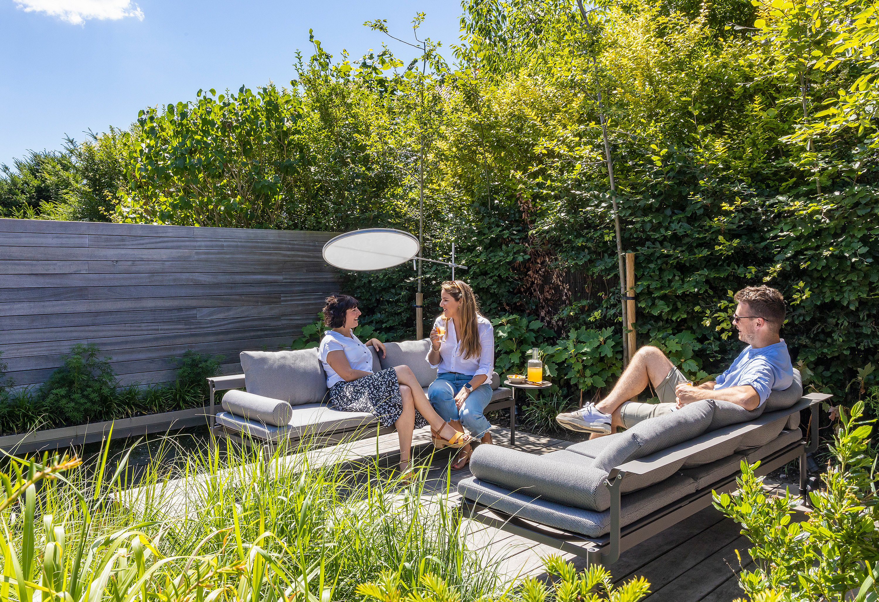 Three adults sitting and reclining on gray Sol+Luna loungers beside a natural pool, with a round white parasol and dense green planting behind them.