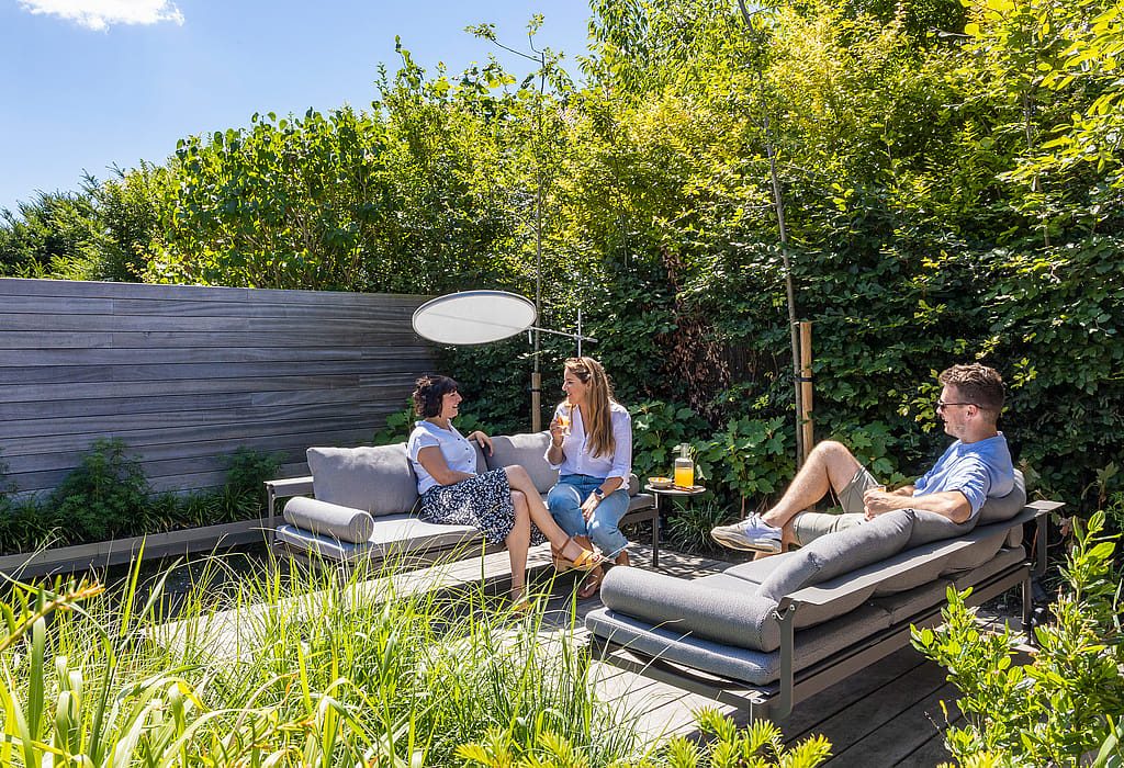 Three adults sitting and reclining on gray Sol+Luna loungers beside a natural pool, with a round white parasol and dense green planting behind them.