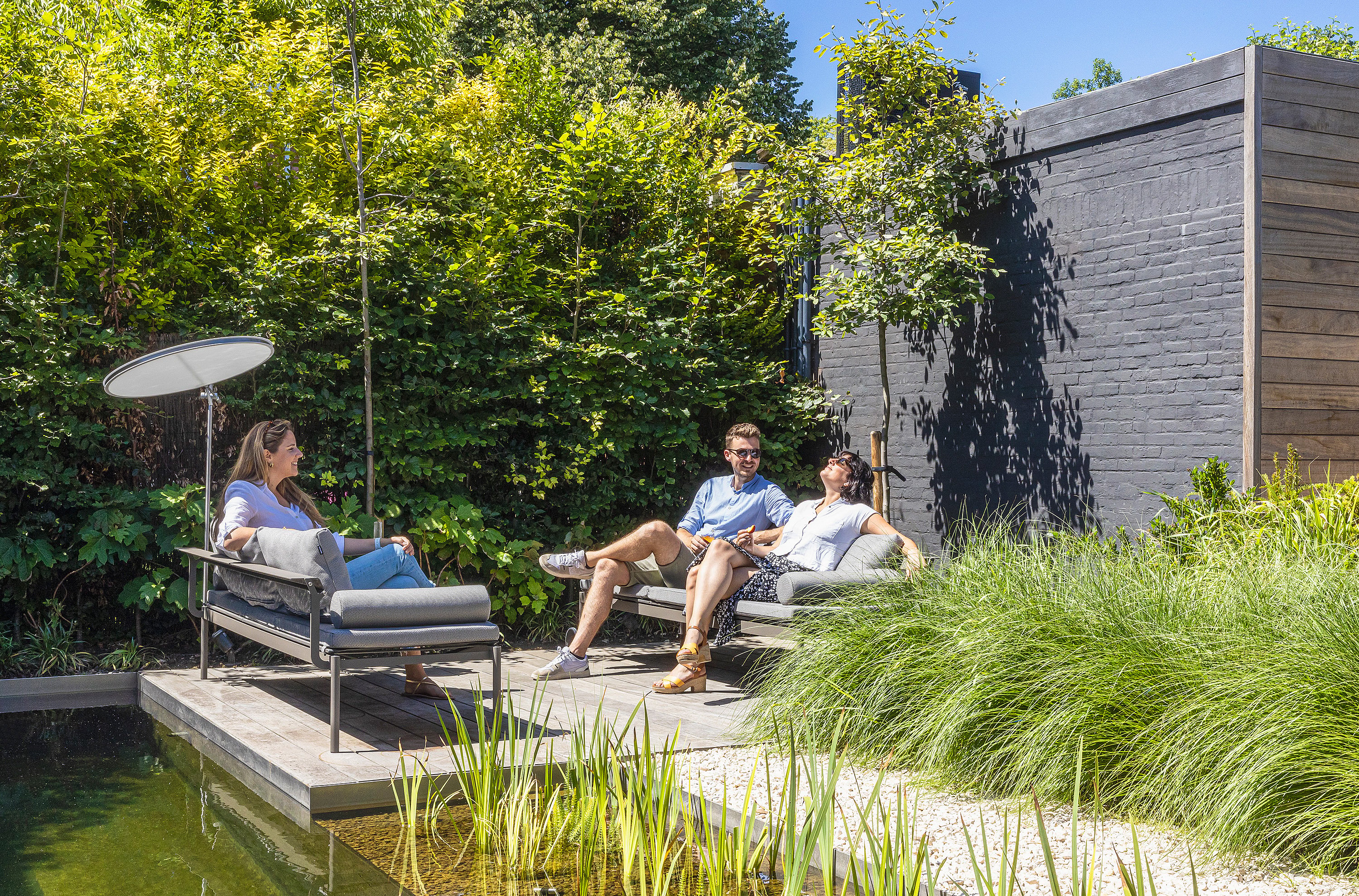 Drei Erwachsene entspannen auf grauen Sol+Luna-Liegen auf einer Holzterrasse neben einem natürlichen Swimmingpool, umgeben von üppigem Grün und einer dunklen Gartenmauer aus Holz.