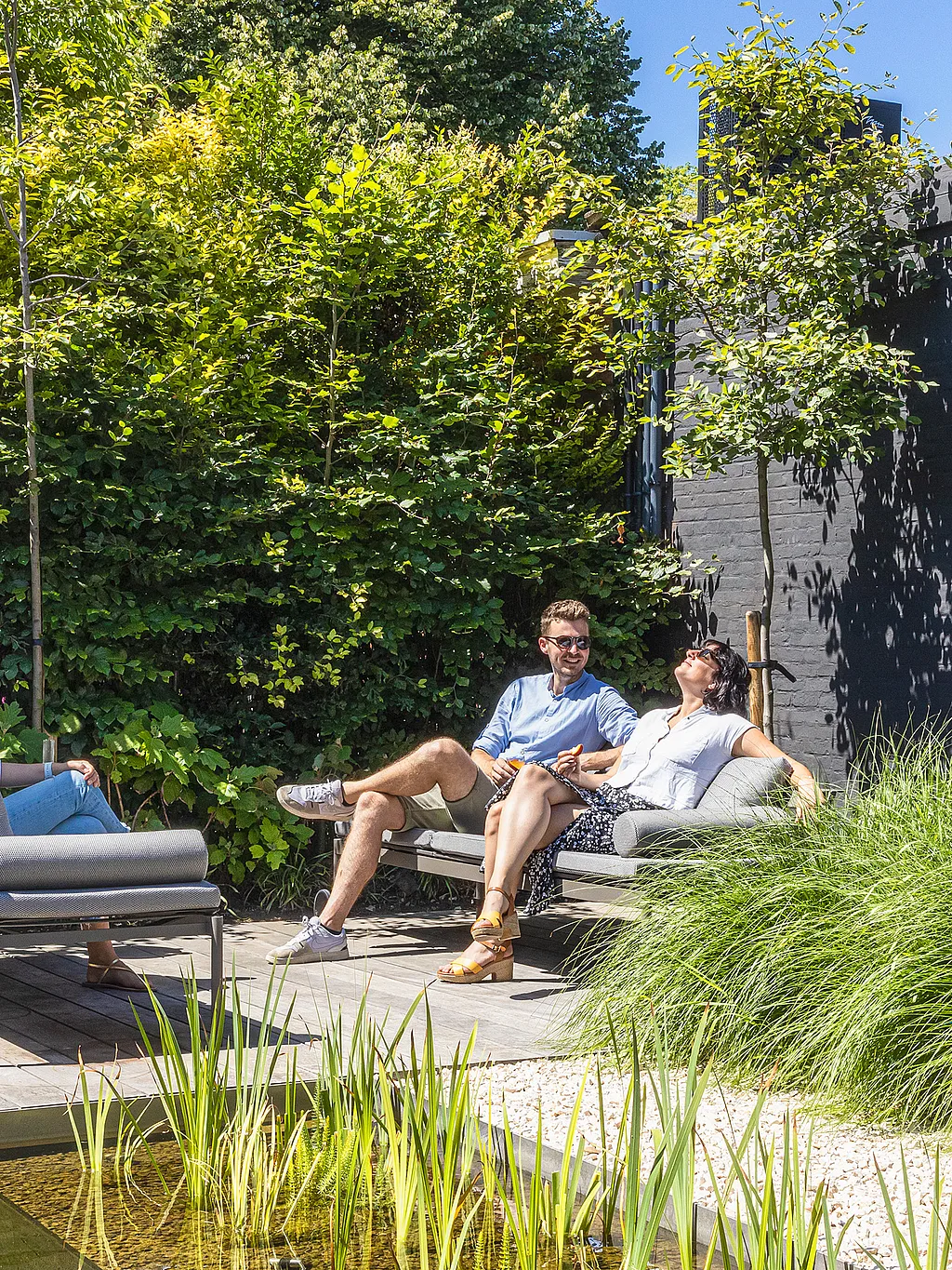 Drei Erwachsene entspannen auf grauen Sol+Luna-Liegen auf einer Holzterrasse neben einem natürlichen Swimmingpool, umgeben von üppigem Grün und einer dunklen Gartenmauer aus Holz.