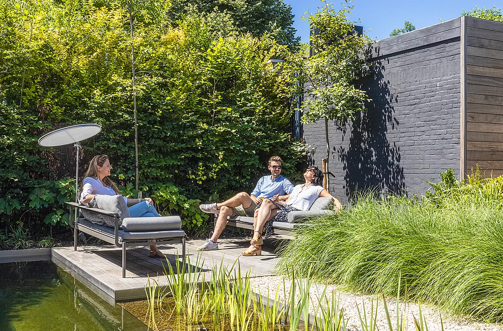 Three adults relaxing on gray Sol+Luna loungers on a wooden deck beside a natural swimming pool, surrounded by lush greenery and a dark timber garden wall.