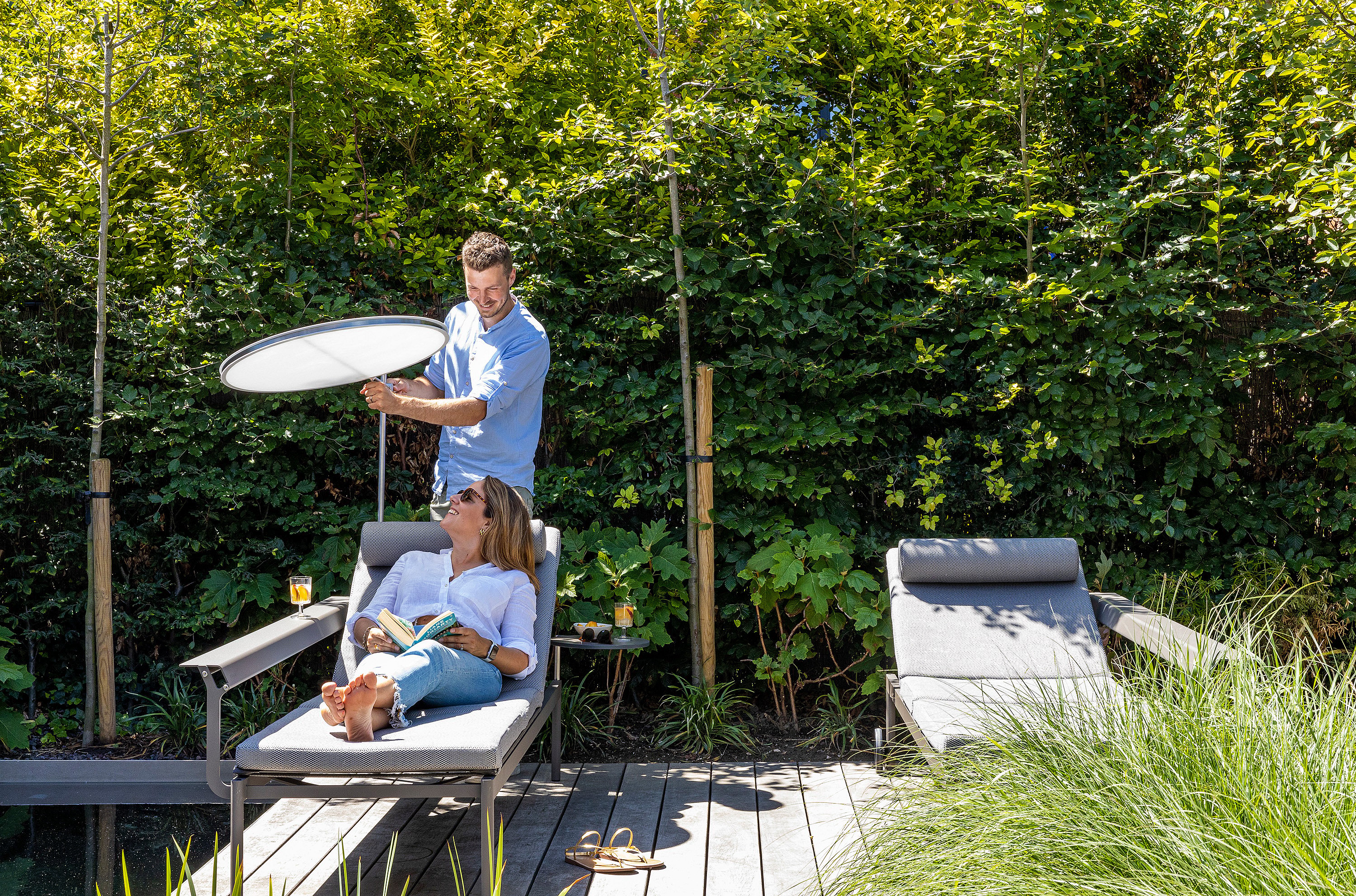 Man adjusting a round white parasol while a woman reclines on a gray lounger beside a natural swimming pool, with a second lounger nearby.