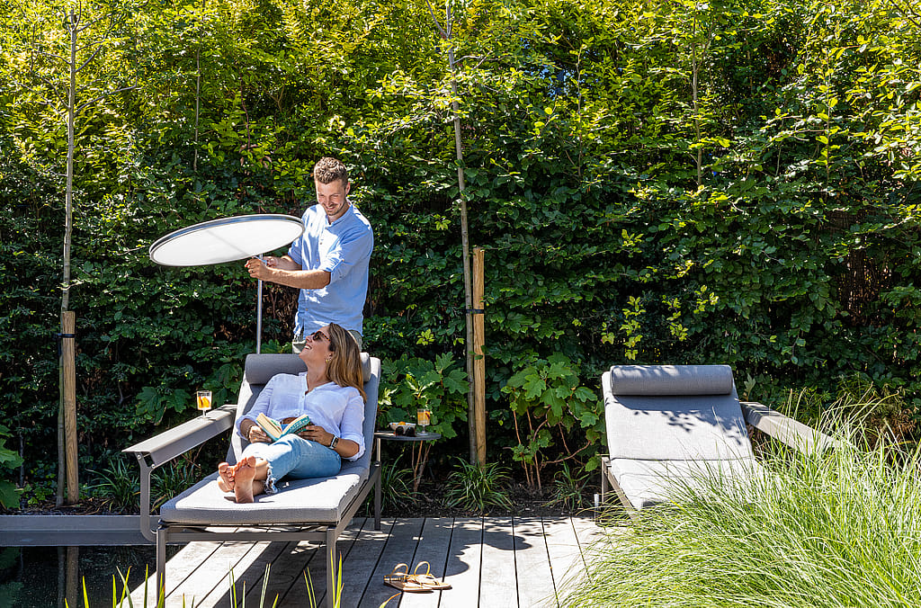 Man adjusting a round white parasol while a woman reclines on a gray lounger beside a natural swimming pool, with a second lounger nearby.
