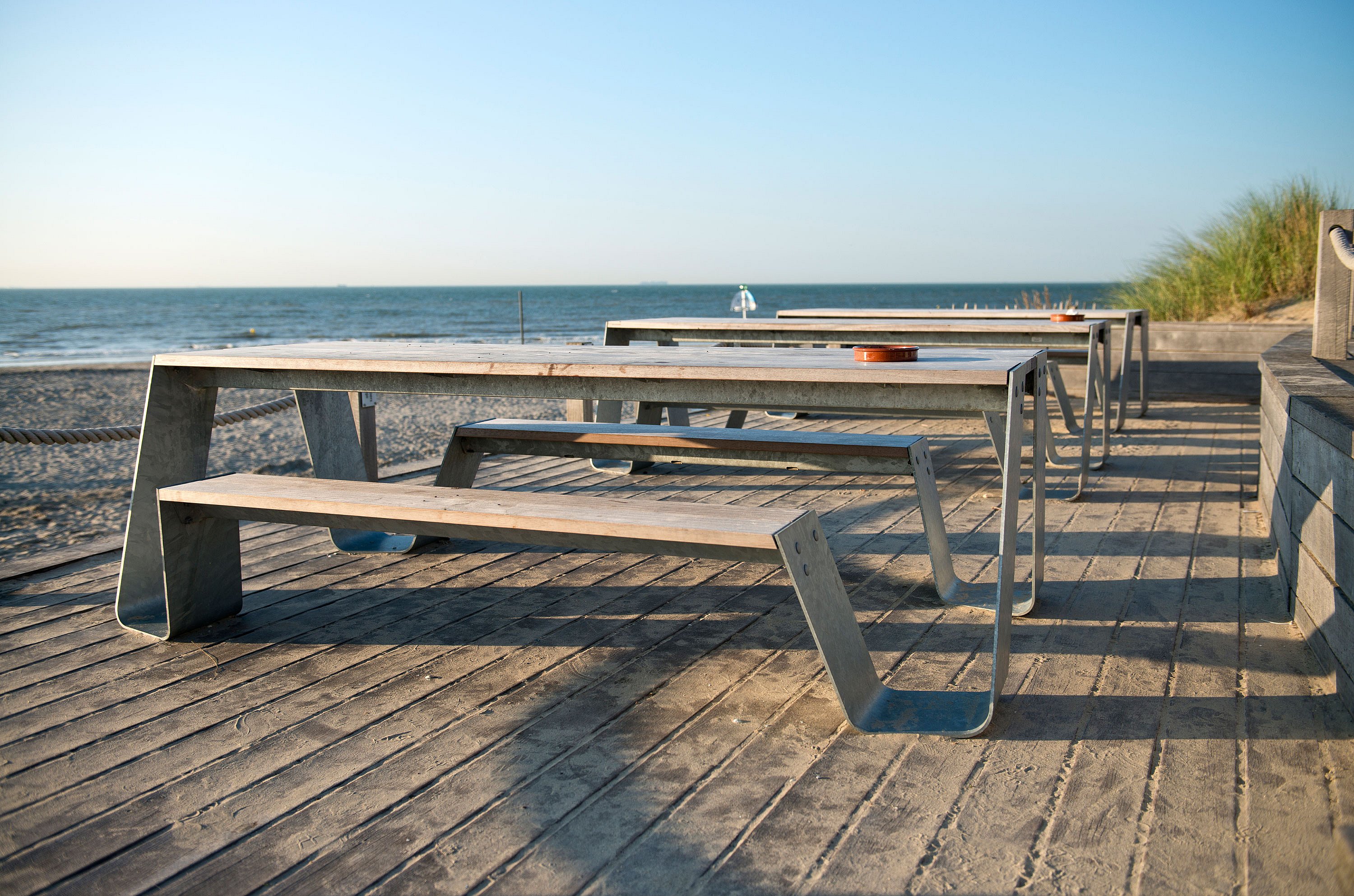   Rangée de tables de pique-nique Hopper avec plateaux en bois et pieds galvanisés sur une terrasse de plage, face à la mer sous un ciel bleu clair.