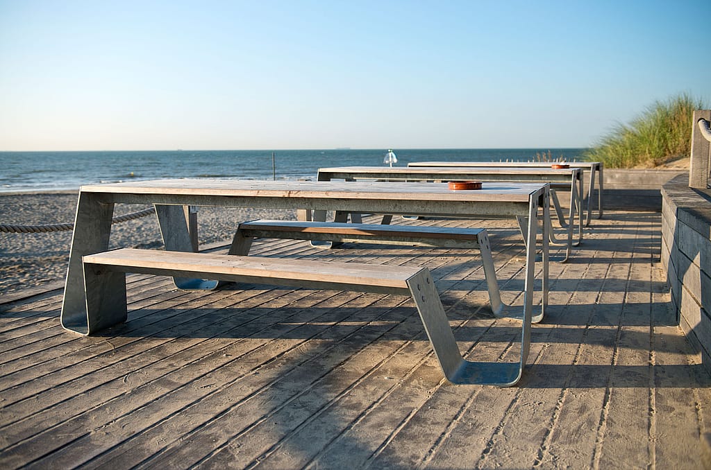 Rangée de tables de pique-nique Hopper avec plateaux en bois et pieds galvanisés sur une terrasse de plage, face à la mer sous un ciel bleu clair.