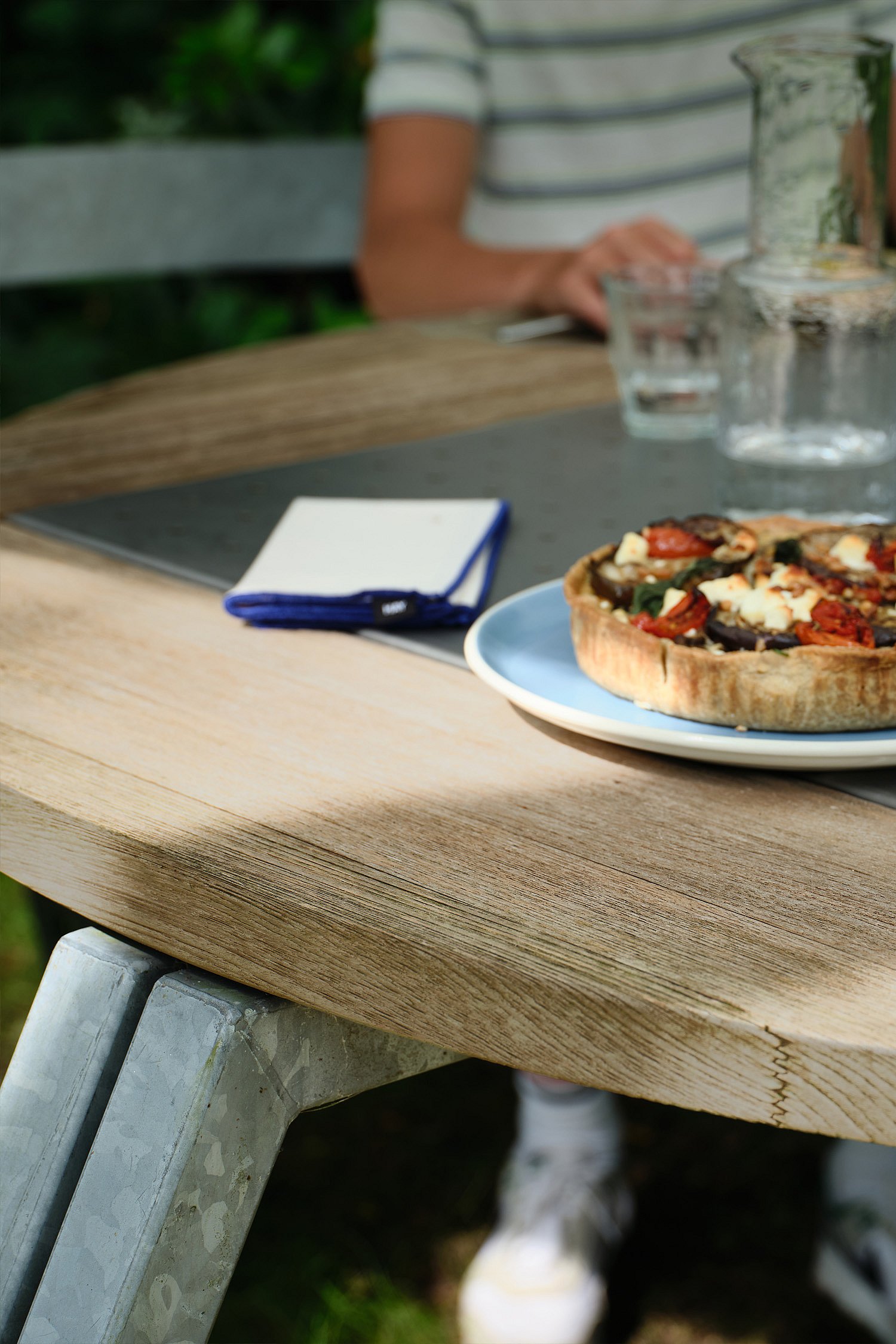 Close-up of a wooden round tabletop with a savory tart on a plate, glasses, and a folded napkin, with a seated person softly blurred behind.